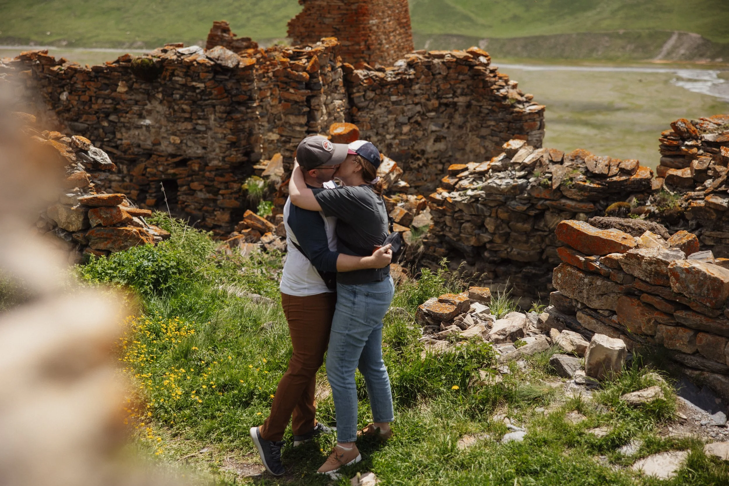 A couple sharing a kiss among ruins of old stone walls in a green scenic landscape.