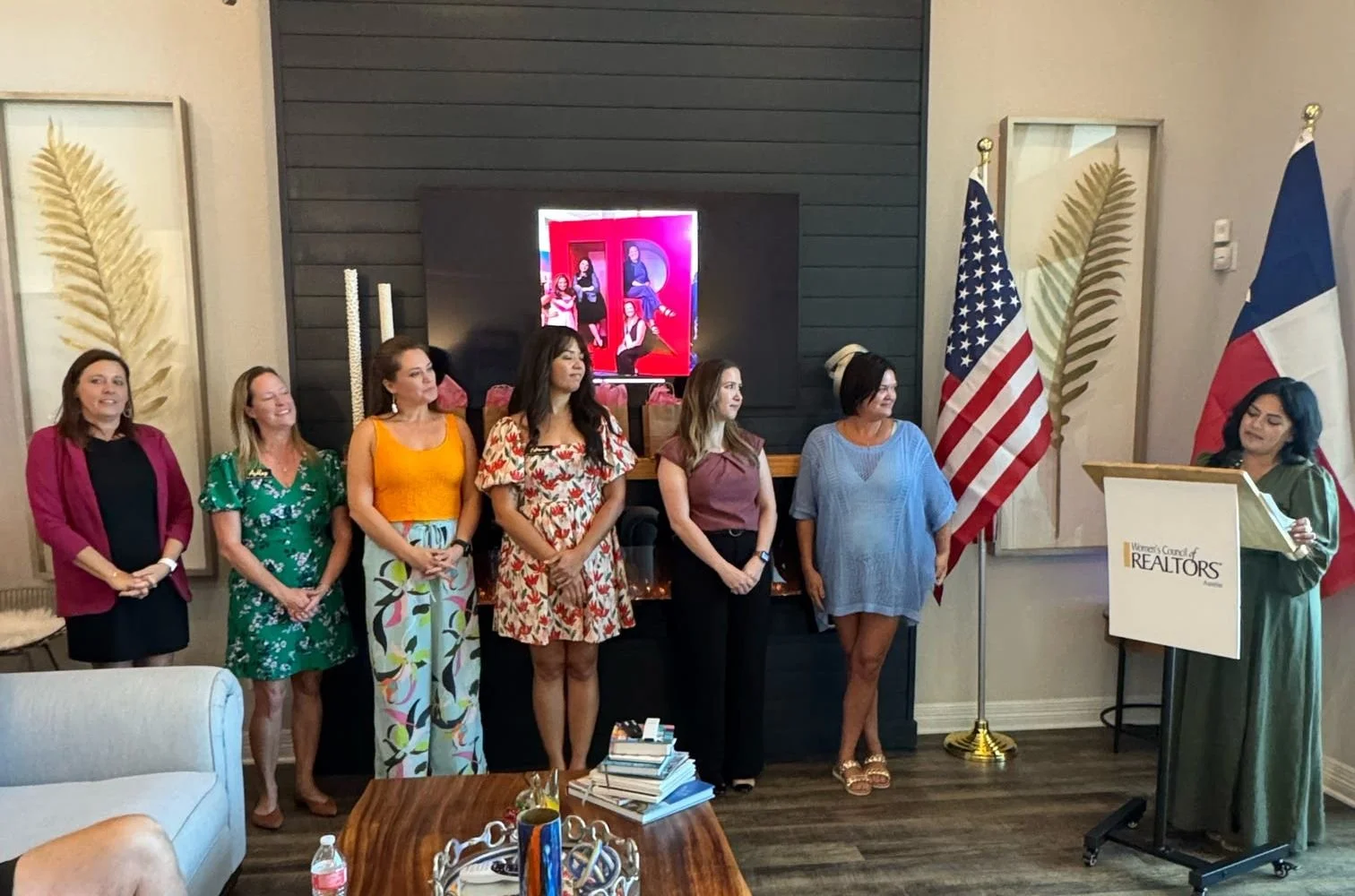 A woman at a podium speaking to a group of women standing in a line. The women are dressed in colorful dresses and skirts, and they appear to be at an indoor event with American and Texas flags displayed behind them. The room has decorative artwork a