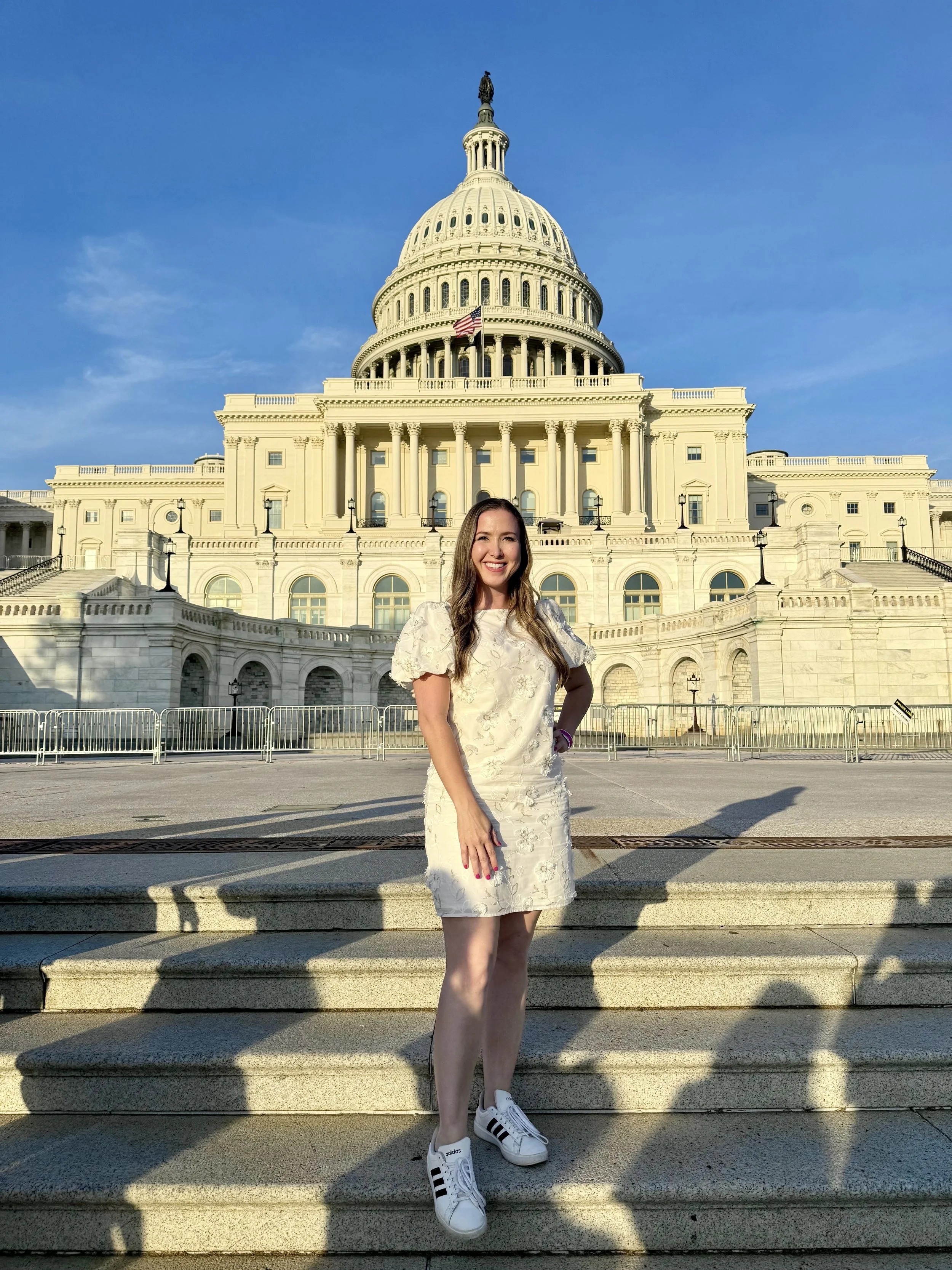 A woman with long hair standing on steps in front of the U.S. Capitol building, smiling at the camera, wearing a light-colored dress and white sneakers.