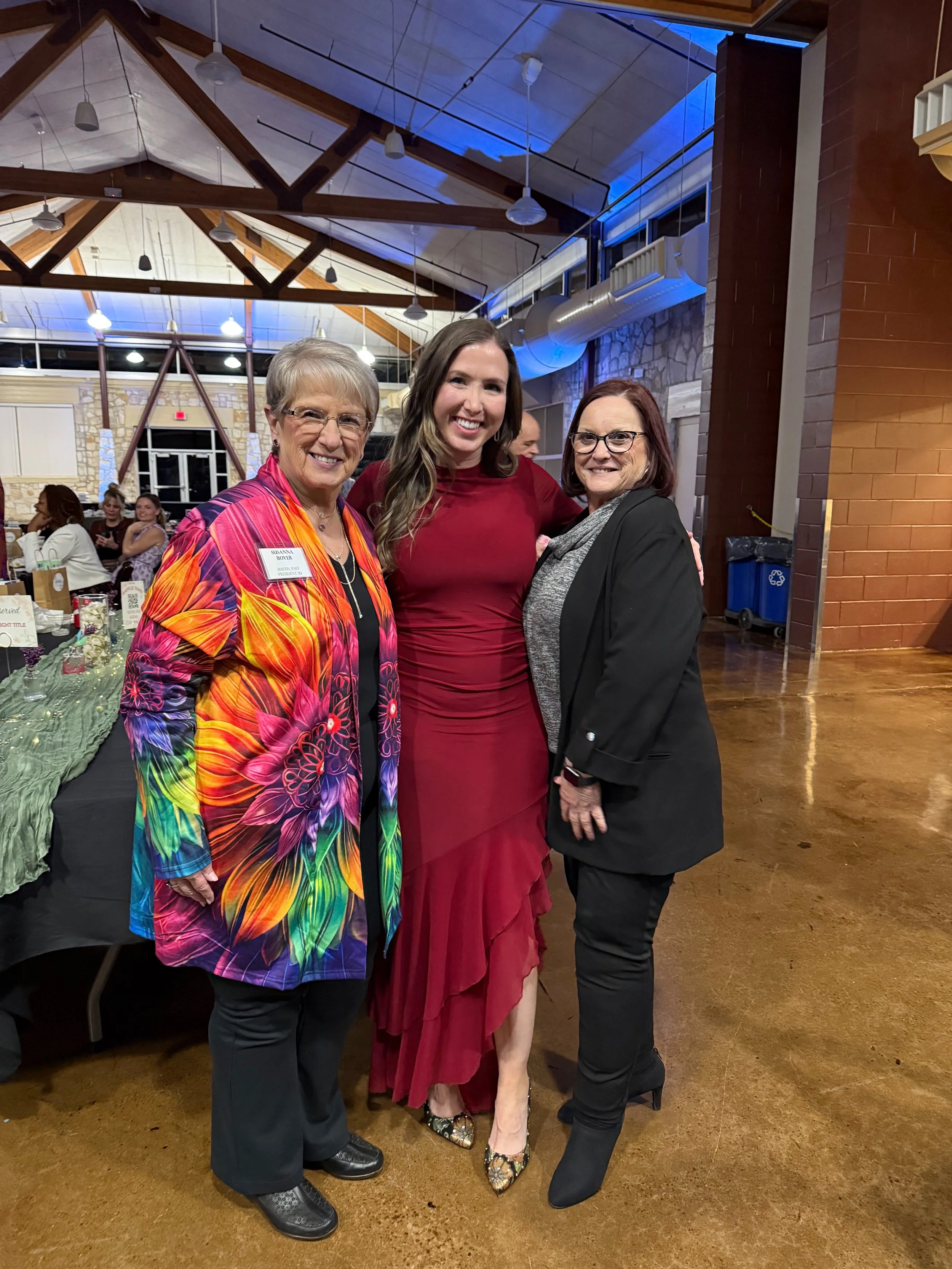 Three women standing together indoors, smiling at the camera, with tables and chairs and other people in the background.
