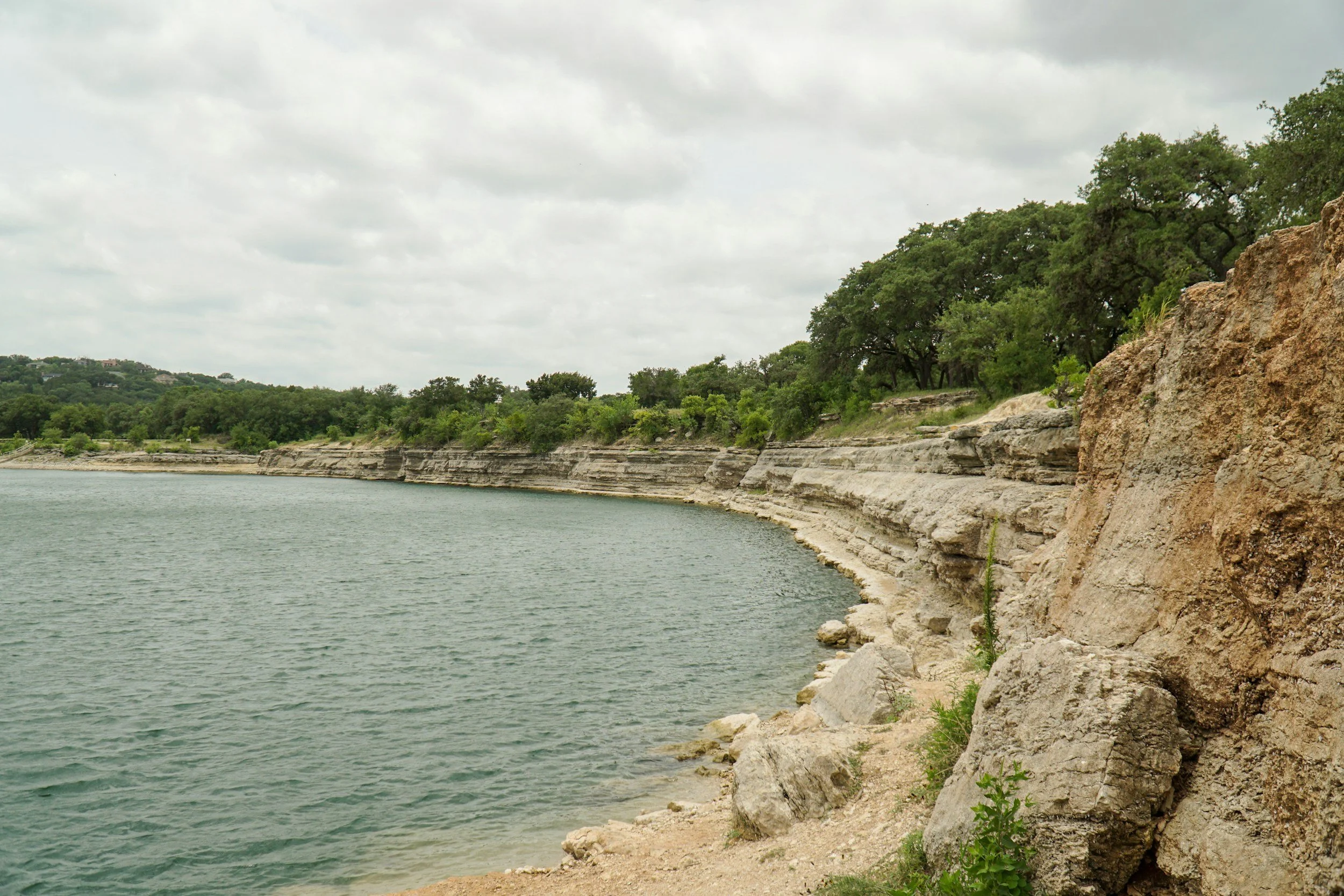 Photo of a river or lake with layered limestone cliffs and green trees on the shoreline under a cloudy sky.