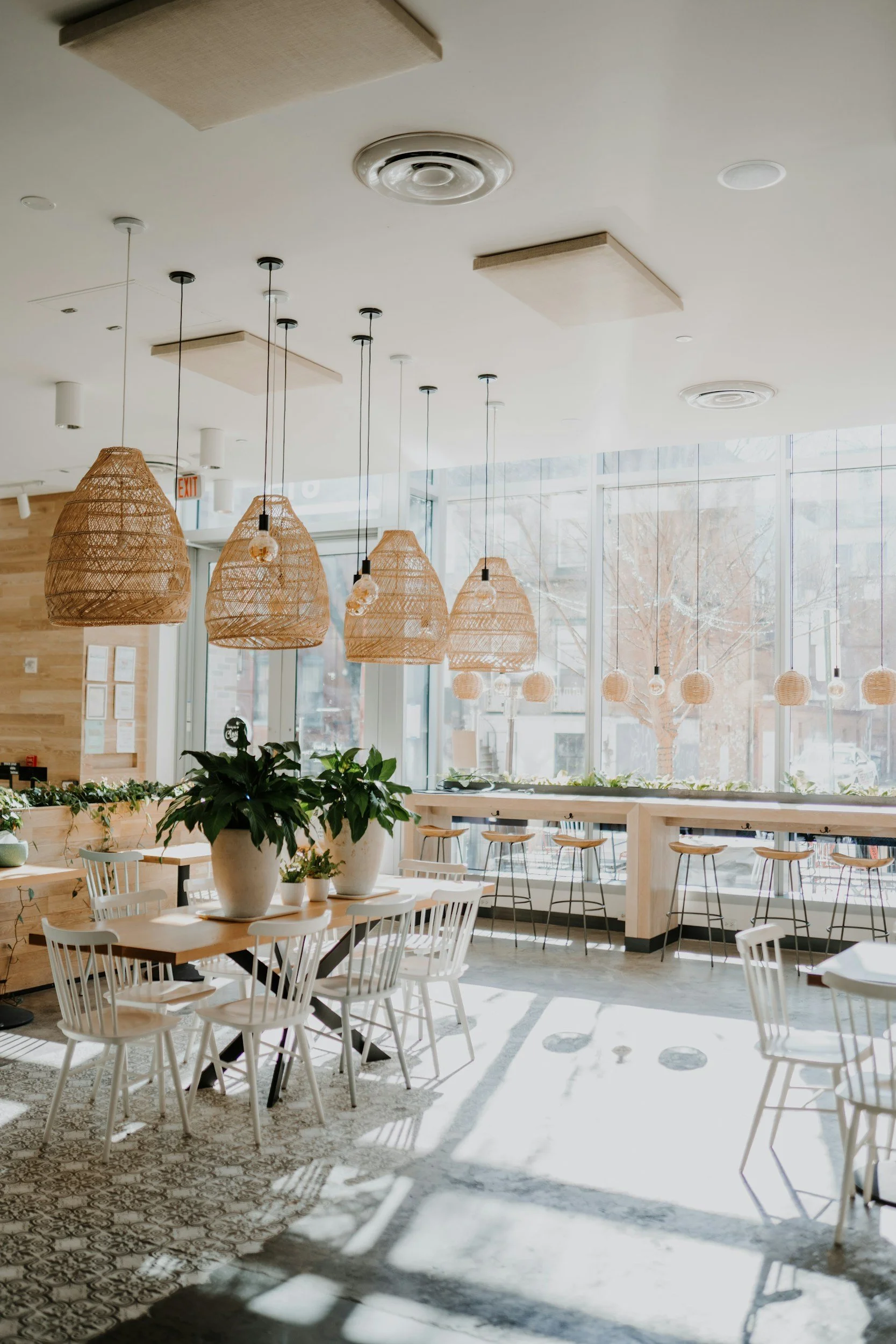 Interior view of a modern, well-lit cafe with white tables, chairs, and large windows. Woven pendant lights hang from the ceiling, and potted plants are on the tables. Sunlight creates shadows on a patterned tile floor.