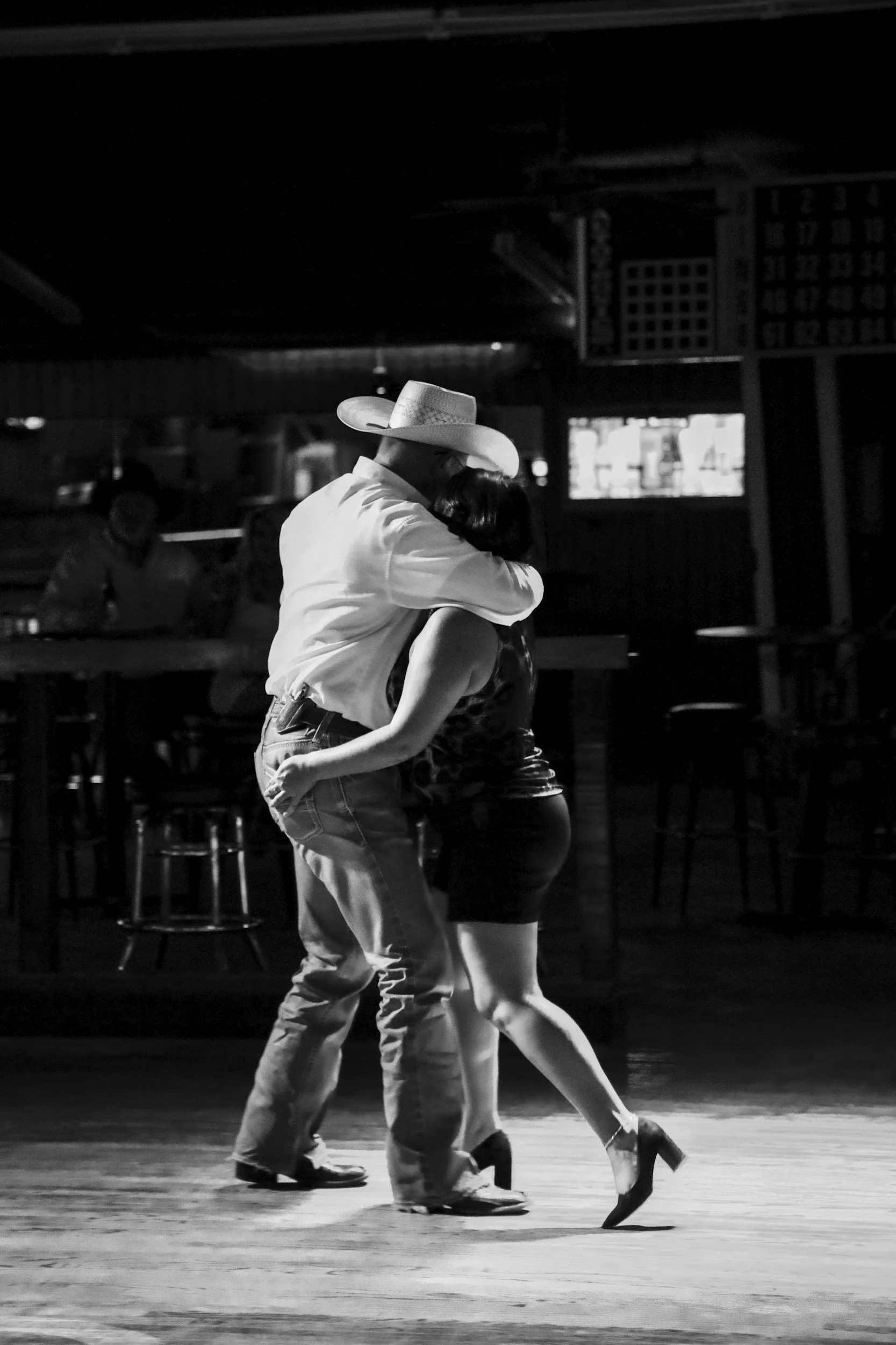 Two people dancing, a man in cowboy hat and a woman in a dress, on a wooden floor in a dimly lit room, with a television in the background.