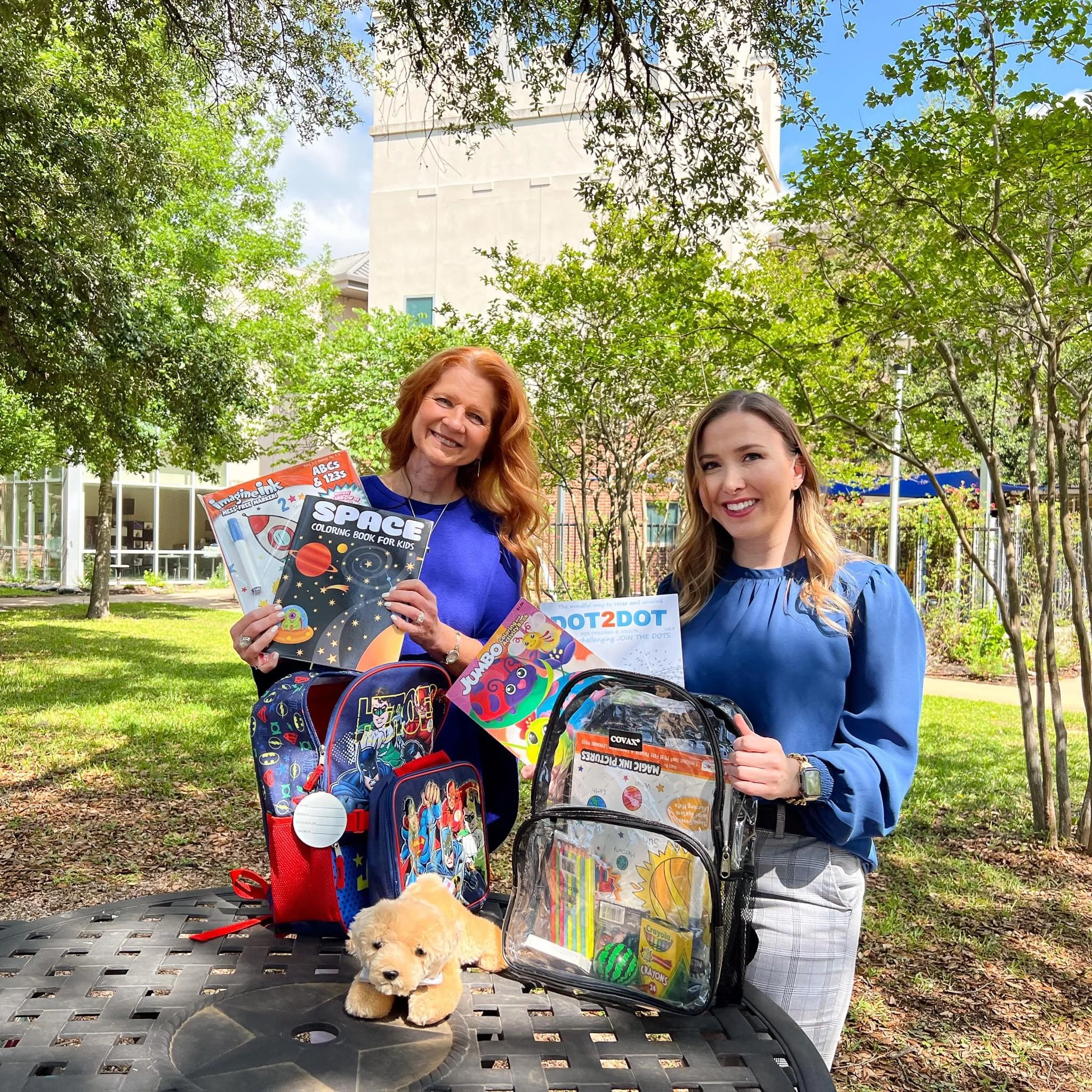 Two women holding school supplies and backpacks outdoors in a park, with a small plush puppy on a table in front of them.