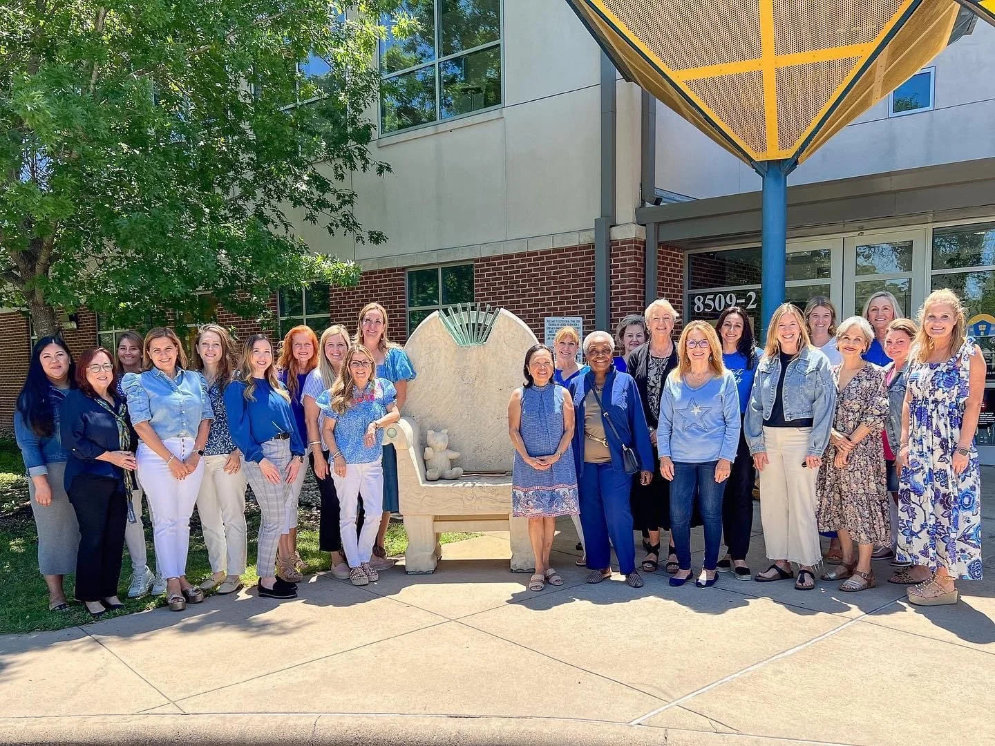 A group of women posing outside in front of a building with large windows and a brick wall, standing next to a decorative sculpture resembling a bed or lounge chair. They are dressed in blue and light colors, with some wearing glasses, and smiling at