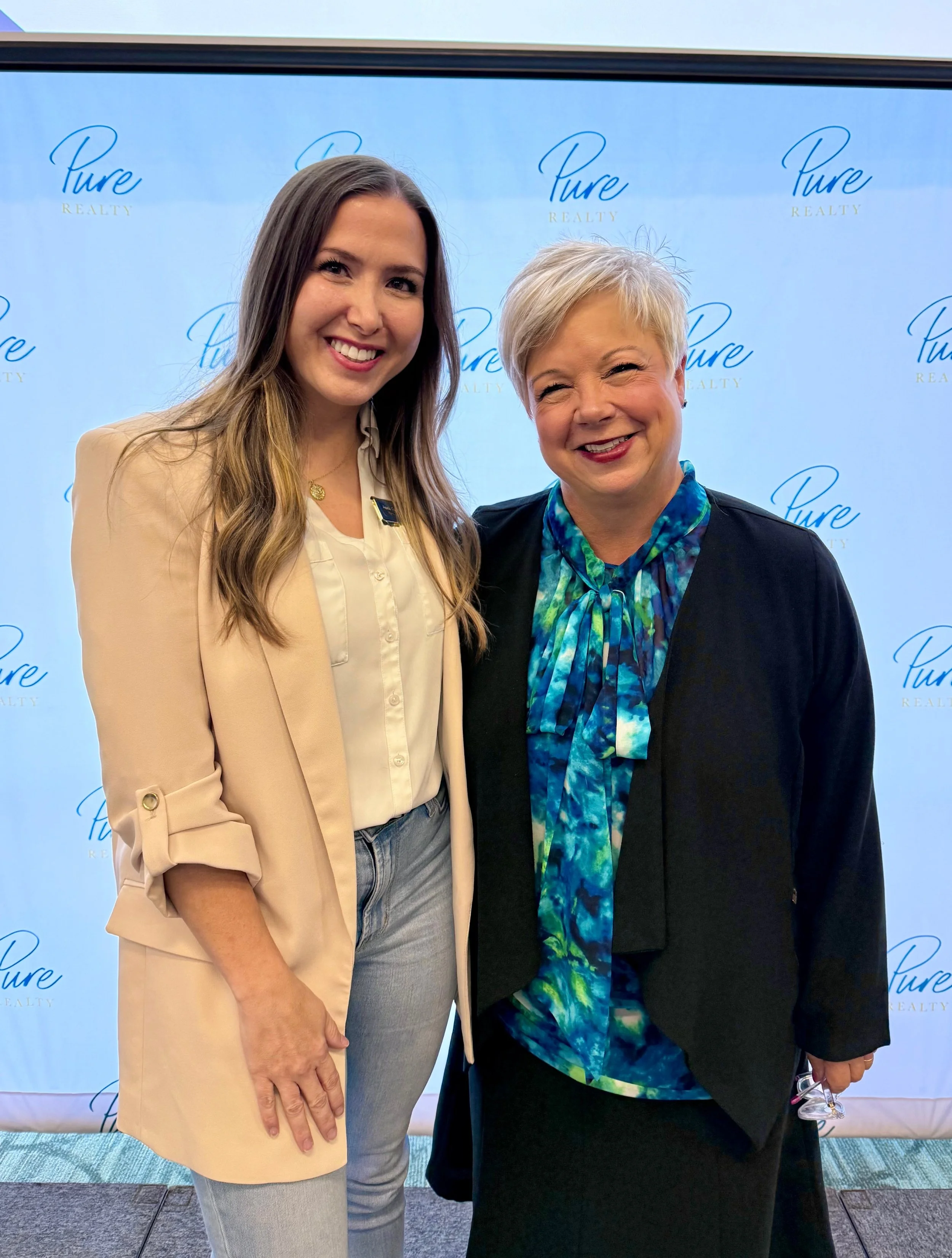 Two women standing side by side, smiling, in front of a blue backdrop with 'Pure Realty' logo repeated, one woman with long blonde hair and light-colored blazer, and the other with short blonde hair and a blue patterned blouse.