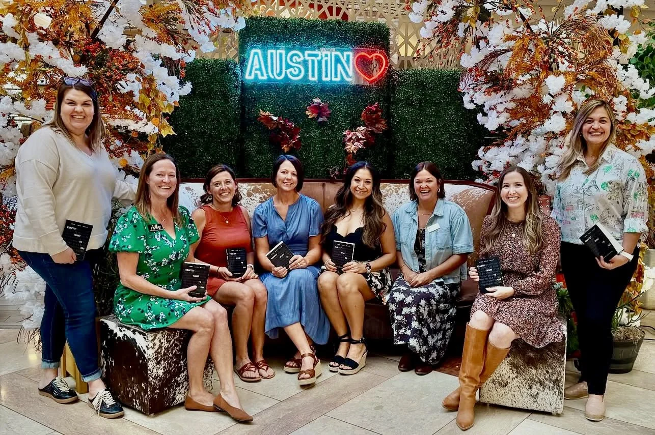 Group of nine women sitting and standing in front of a floral backdrop with the word 'AUSTIN' in neon lights and a red heart, holding books or pamphlets, smiling for the photo.