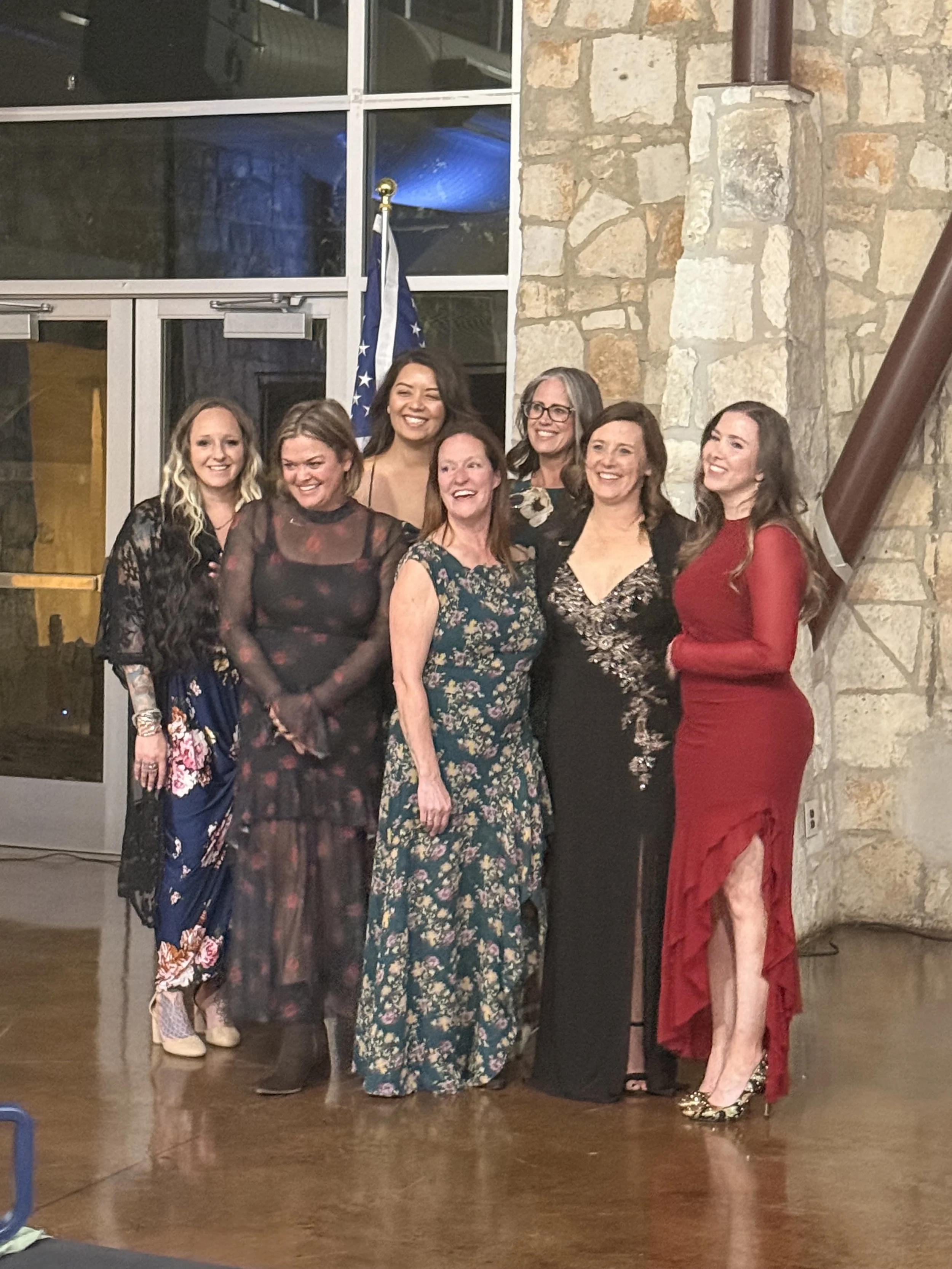 Group of eight women in formal attire standing together at a gathering, smiling for a photo inside a building with stone walls and large glass windows.