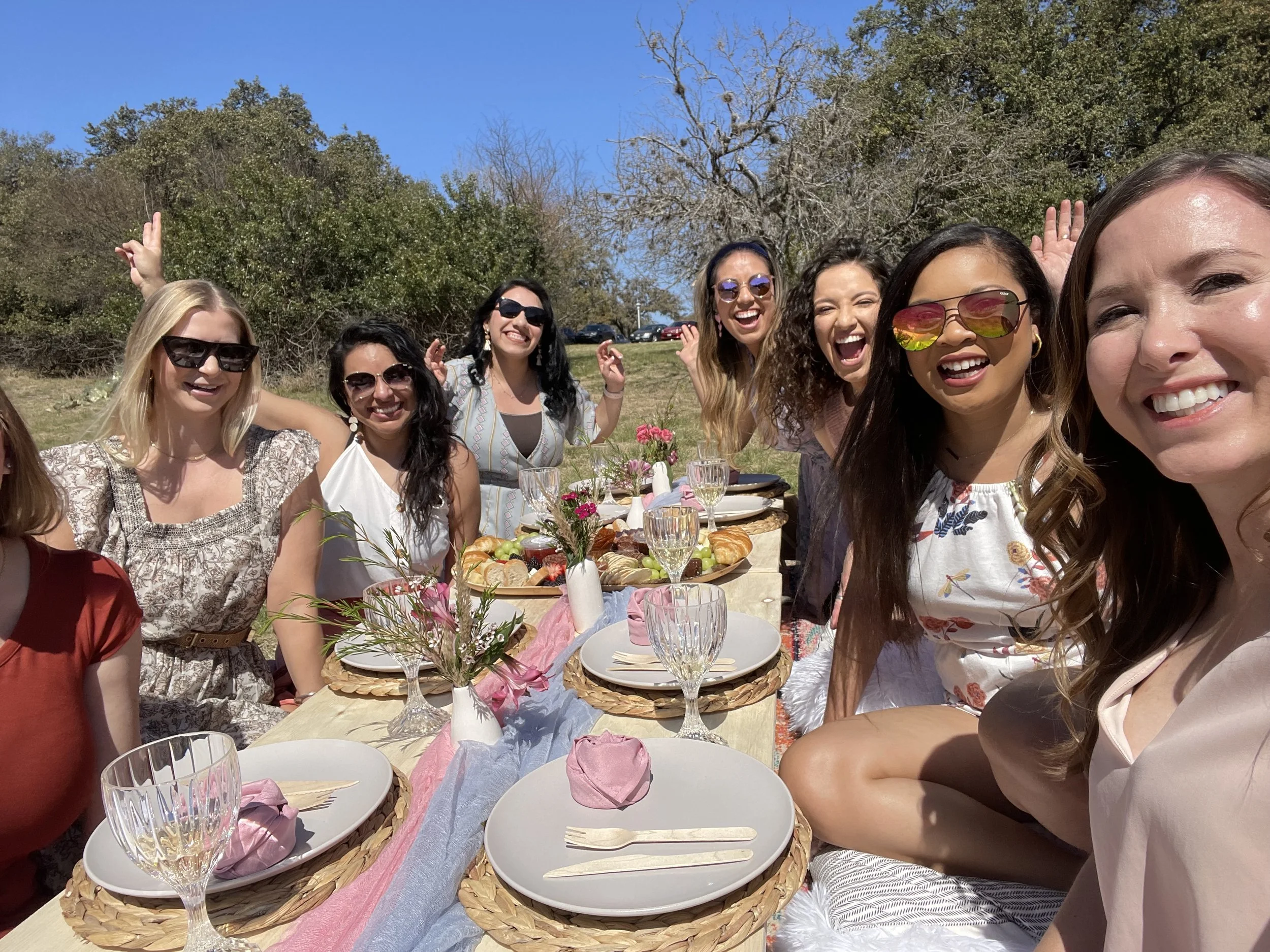 Group of women smiling and posing for a photo at an outdoor table set for a gathering with plates, glasses, and food, on a sunny day in a park with trees in the background.
