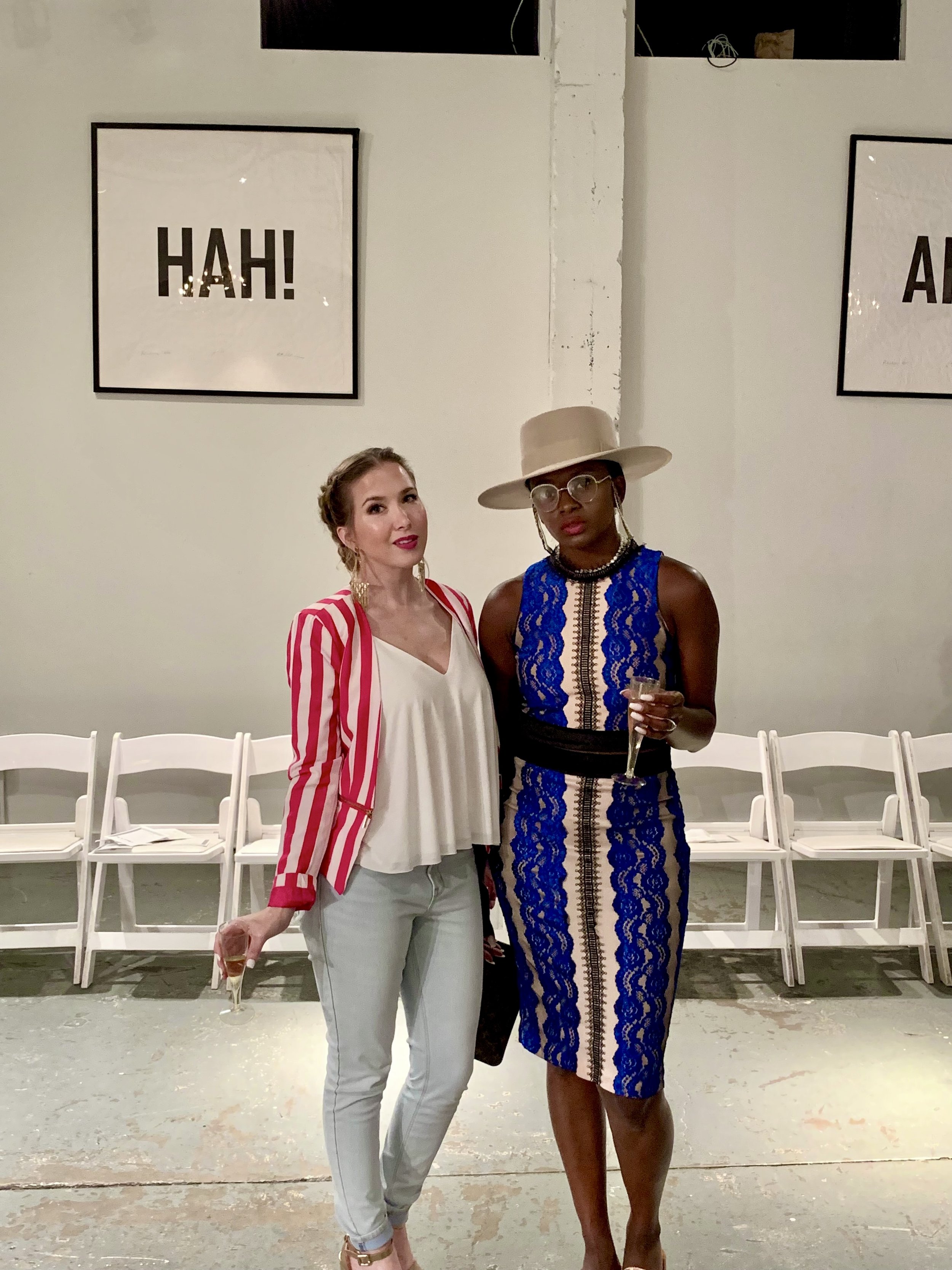 Two women standing side by side, holding glasses of champagne, in an indoor space with simple white chairs arranged in the background and artwork on the wall behind them.
