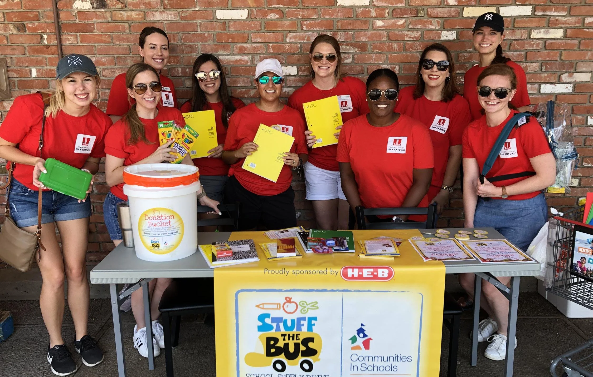 Group of women wearing red shirts and sunglasses standing behind a table with donation materials and a banner. They are smiling and posing in front of a brick wall.