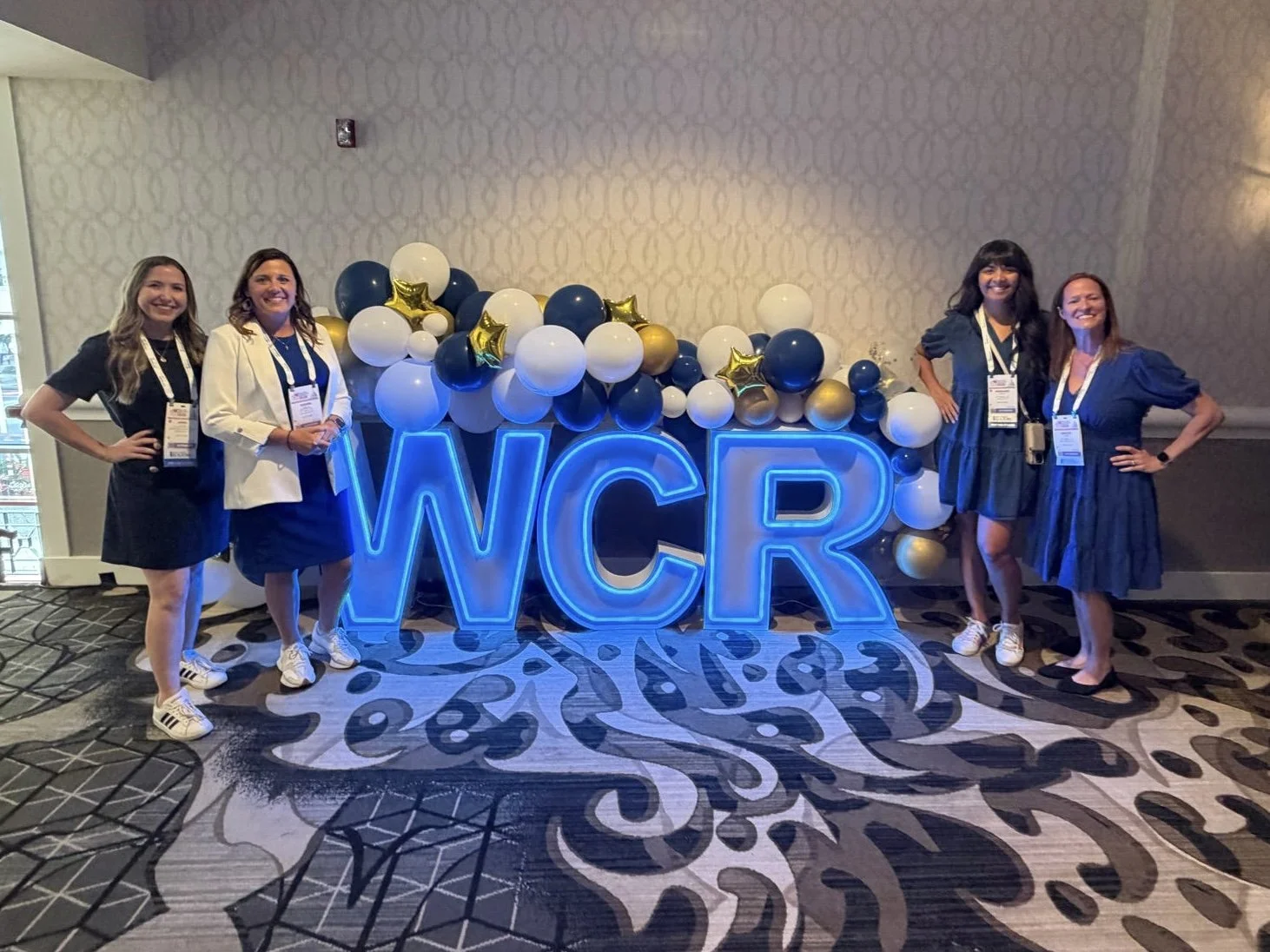 Four women standing in front of a neon sign that reads 'WCR' decorated with balloons in white, blue, black, and gold, with some gold star-shaped balloons, in a conference room.
