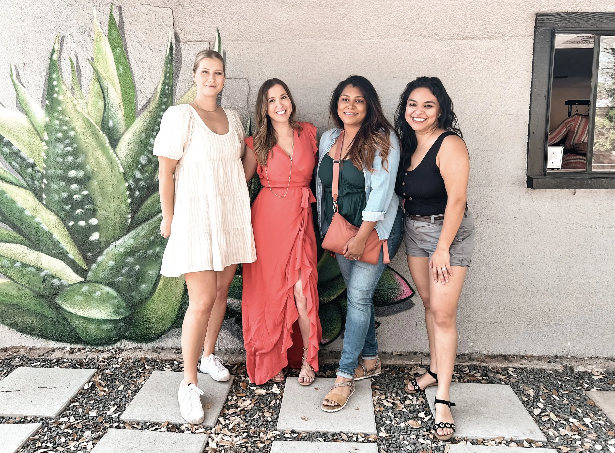 Four women standing on a sidewalk in front of a large painted succulent mural, smiling at the camera.