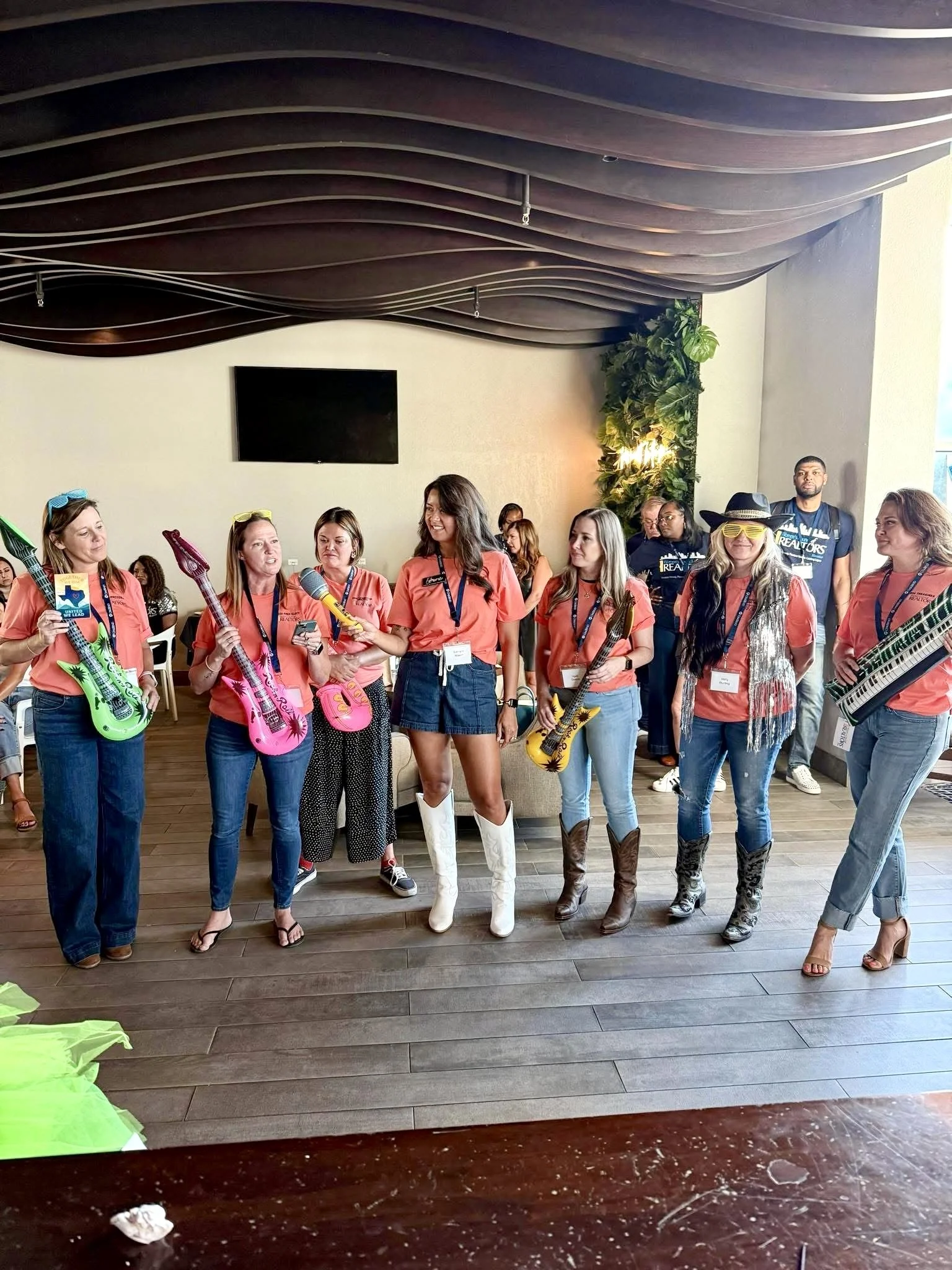 Group of seven women standing indoors, holding colorful toy guitars, wearing orange shirts, jeans or skirts, and some with funky accessories like sunglasses and boots, participating in a group activity or celebration.