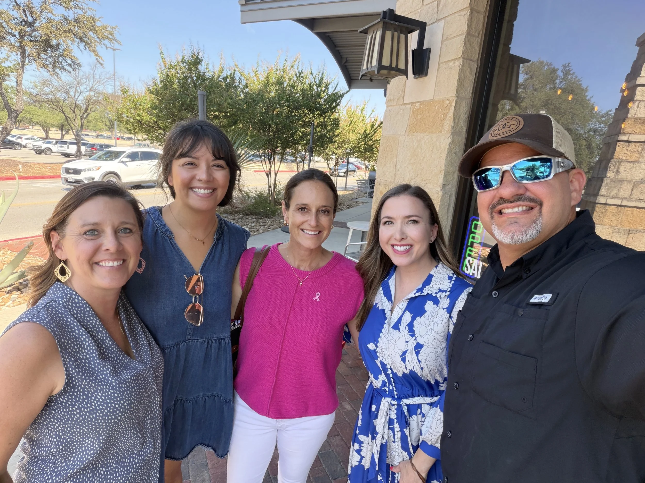 Group of five people smiling in front of a storefront with trees and parked cars in the background.