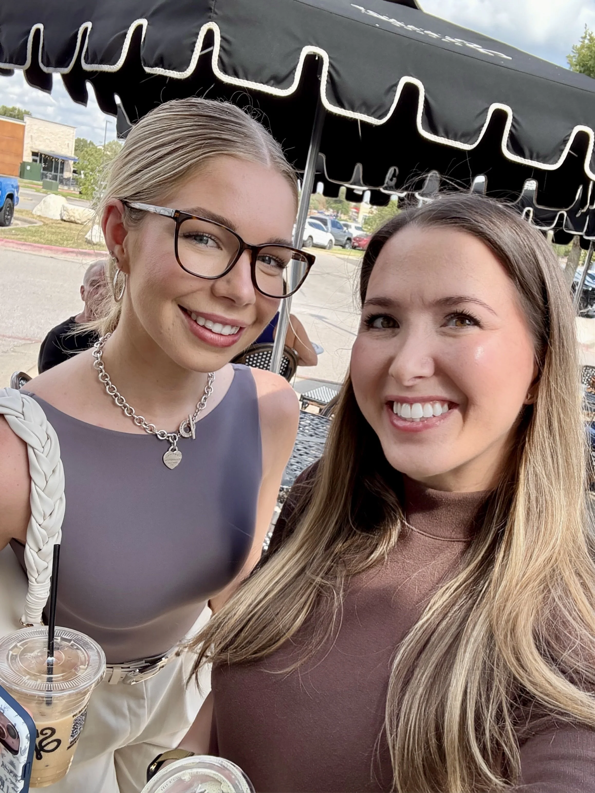 Two women smiling on a patio, with one holding iced coffee drinks, under a black awning on a partly cloudy day.