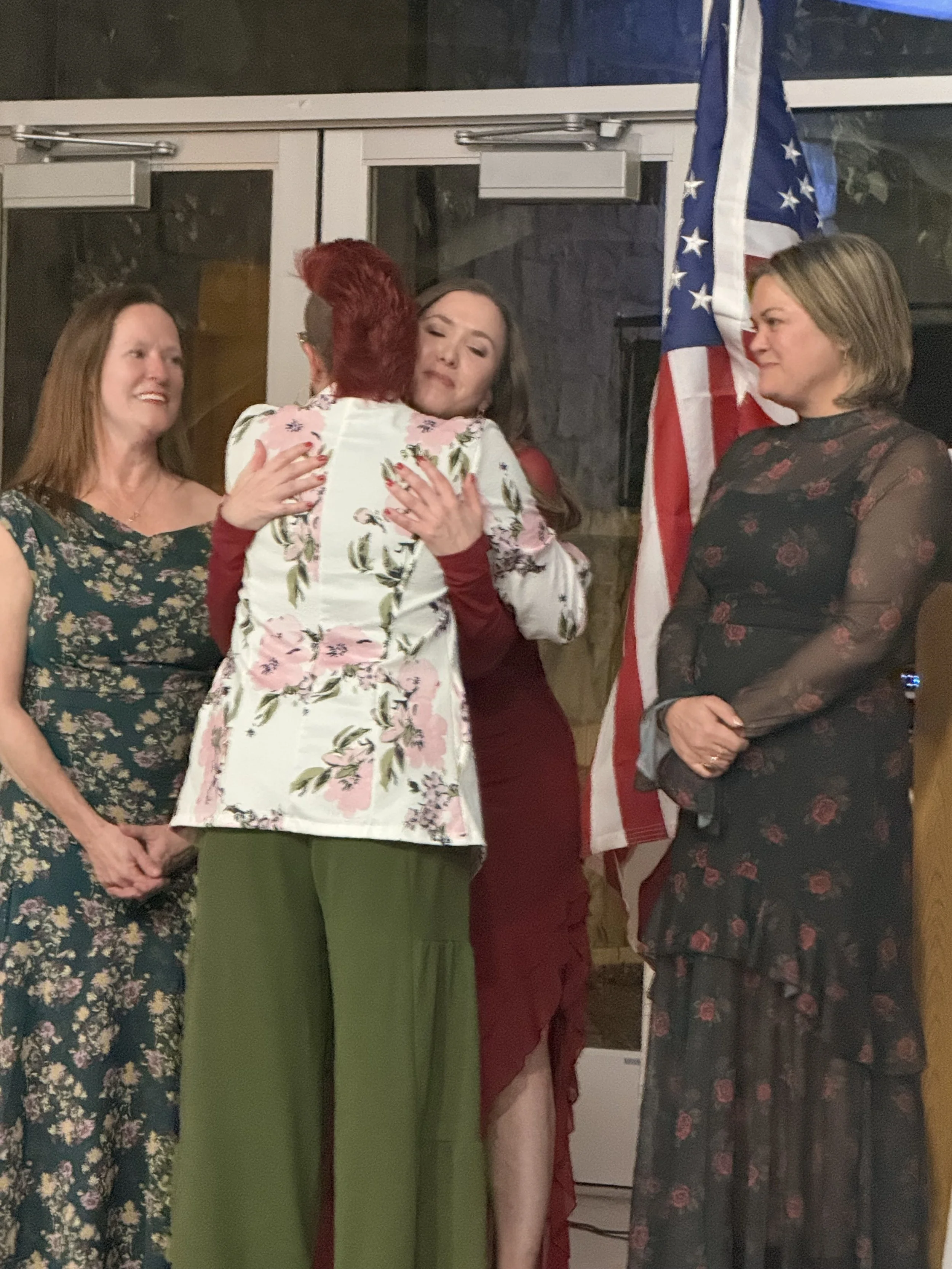 Four women standing together, with one woman hugging another. The woman being hugged is wearing a red dress. An American flag is visible in the background.
