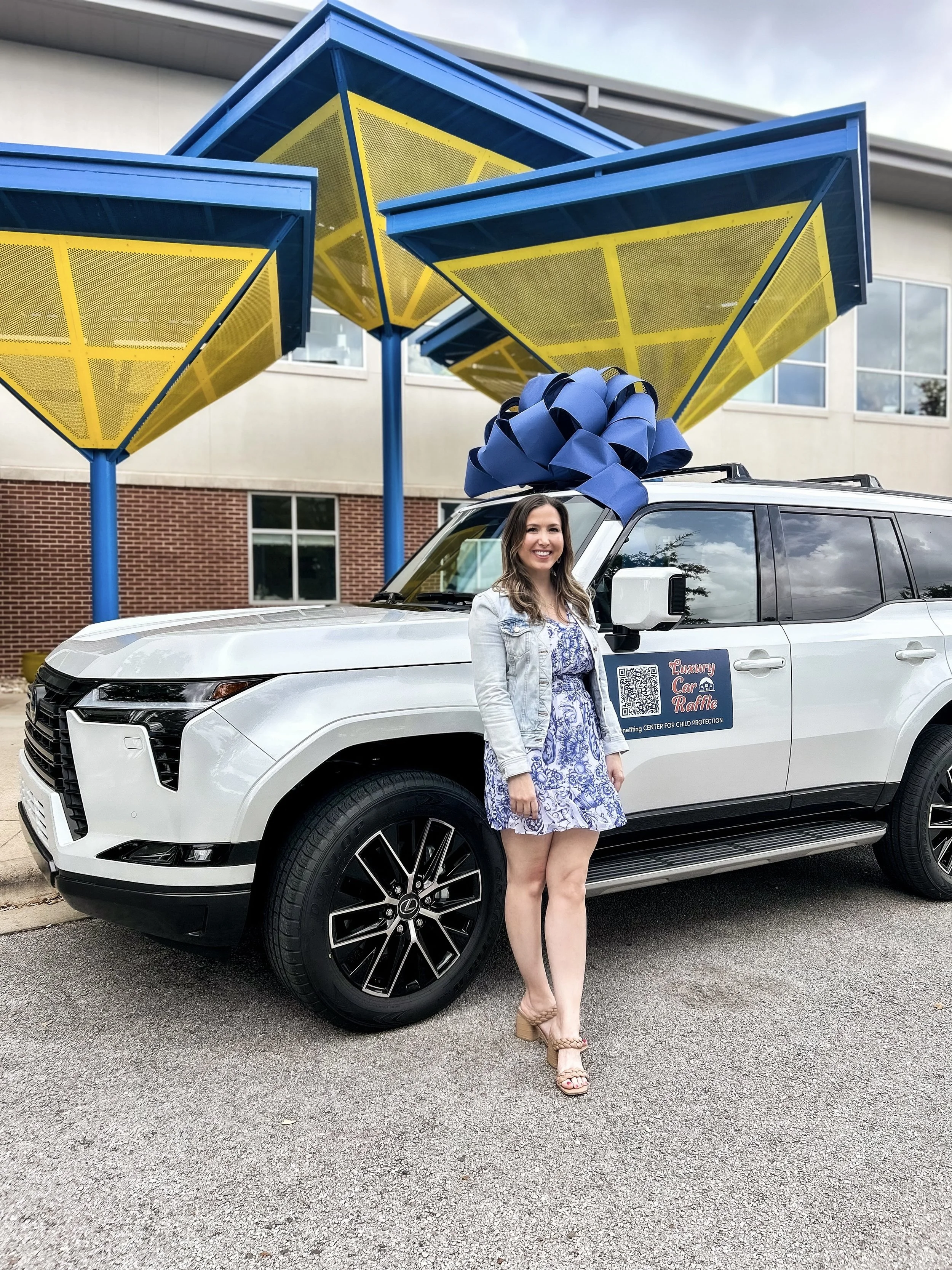Woman standing next to a white luxury SUV with a large blue bow on its roof, parked in front of colorful yellow and blue canopy structures, with a building in the background.