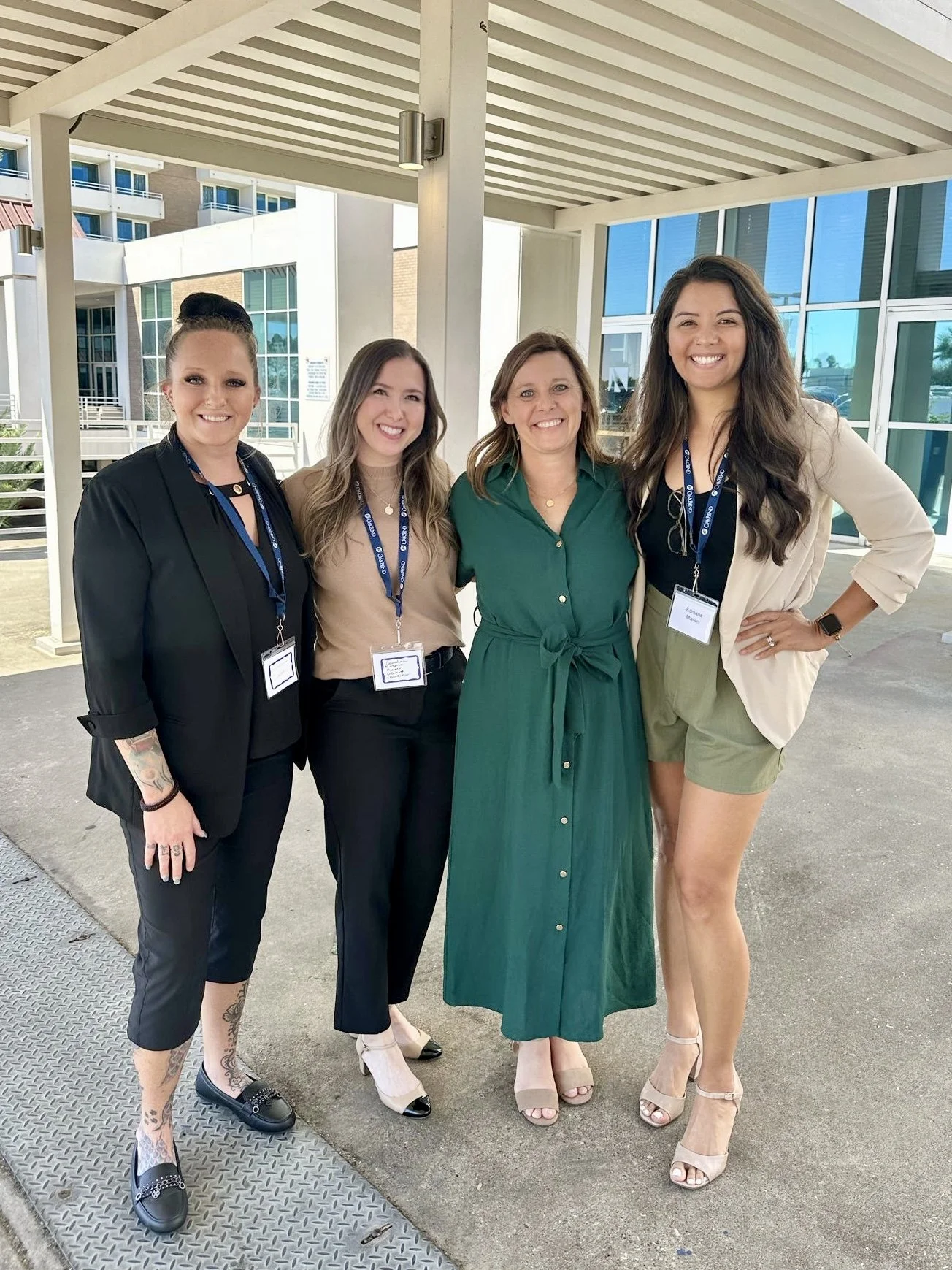 Four women standing together outdoors near a modern building, smiling at the camera. They are wearing business casual and professional attire with name tags and lanyards.