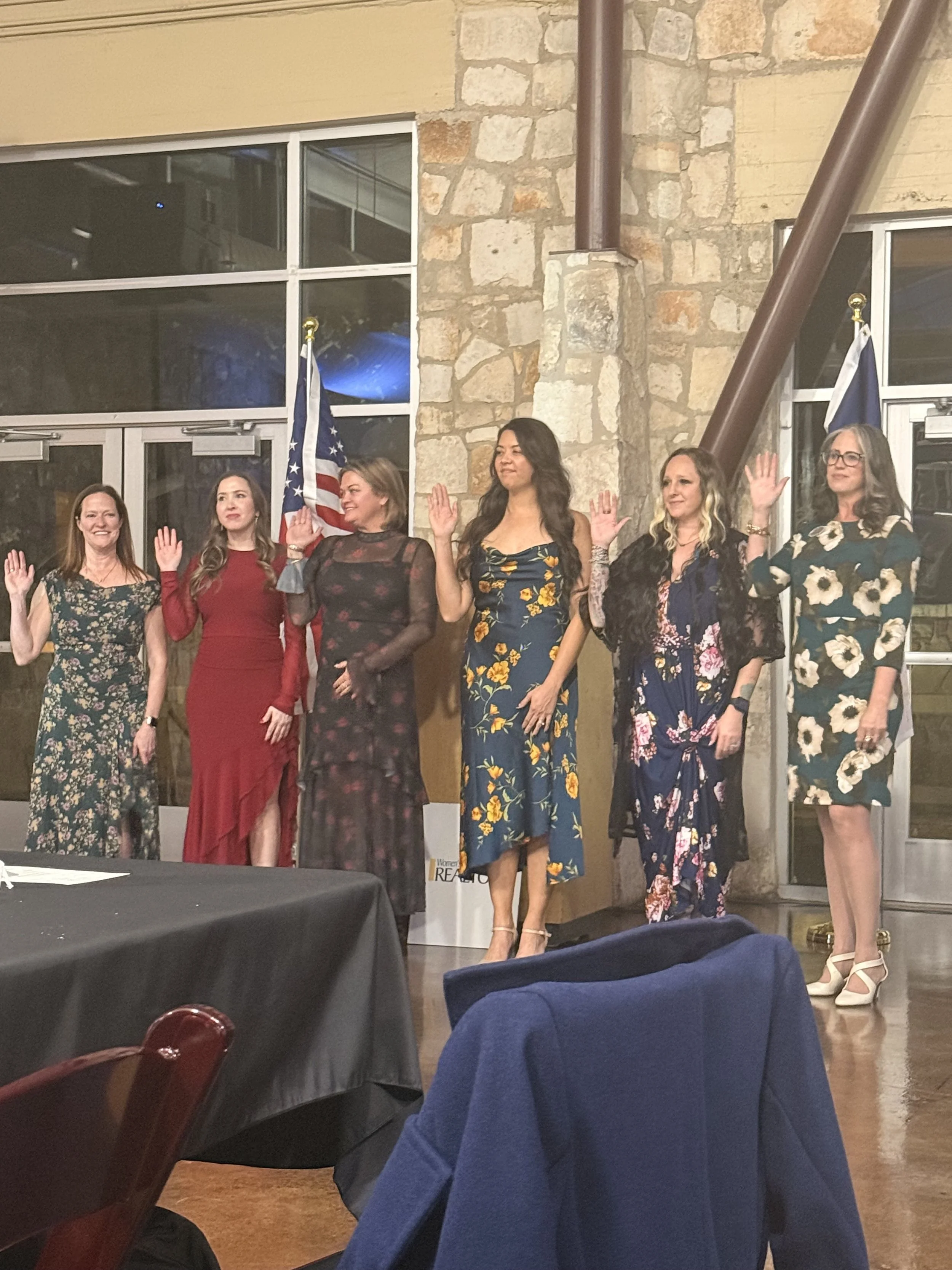 A group of six women standing in a row, raising their right hands. They are dressed in floral and solid-colored dresses, with some wearing high heels, in an indoor setting with stone walls and American flags.