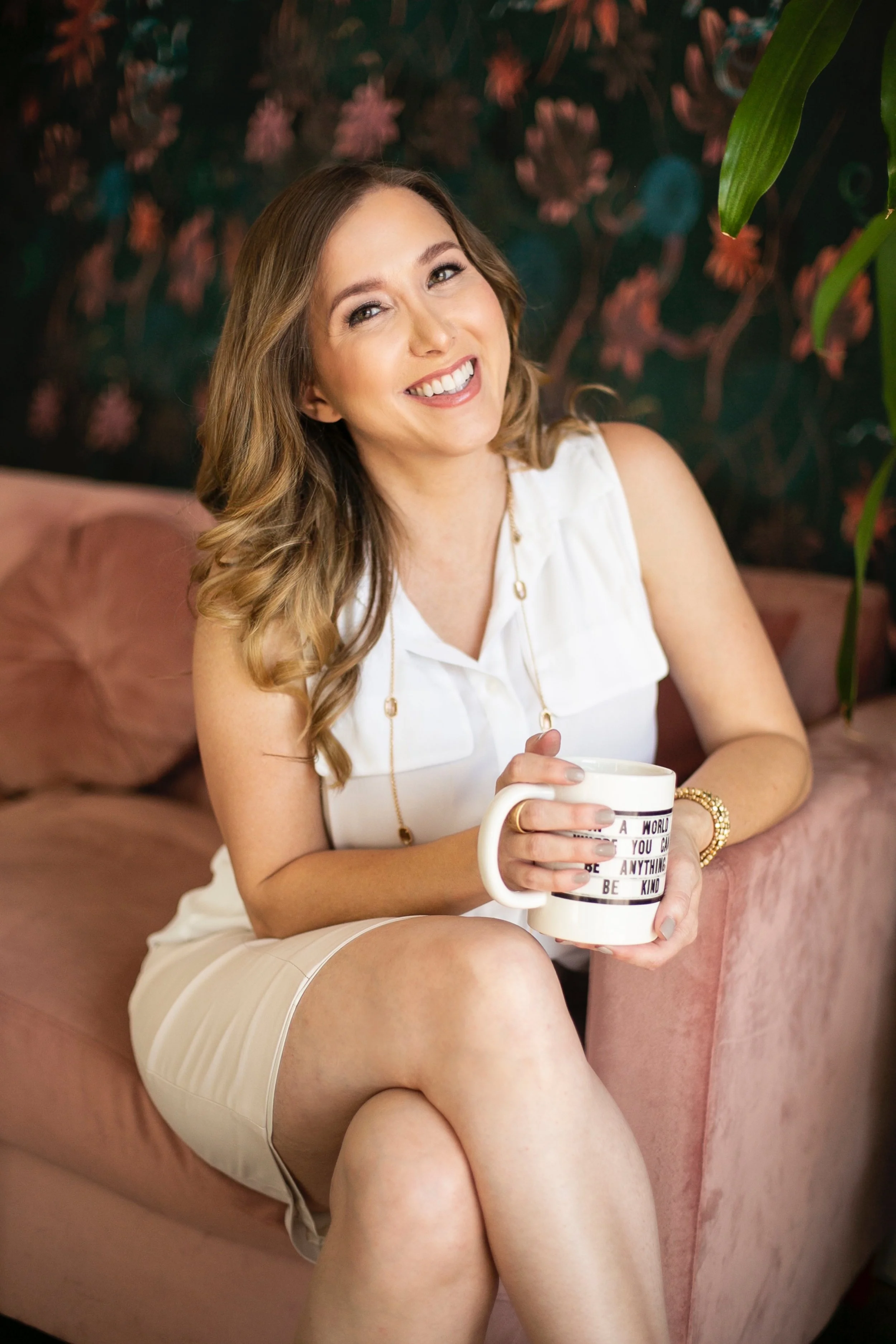 April Guerra with long wavy hair, smiling, sitting on a pink sofa, holding a large coffee mug with text on it. She is wearing a white sleeveless top and beige shorts, against a colorful floral patterned background with greenery.