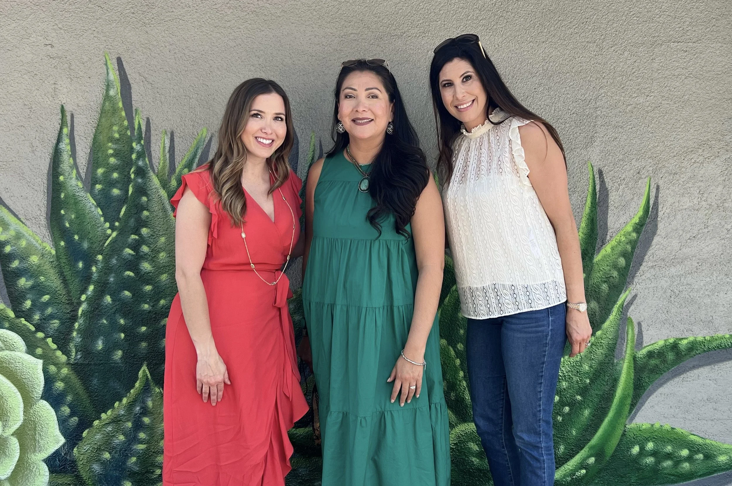Three women standing in front of a painted aloe vera plant on a wall, smiling at the camera, dressed in colorful summer clothing.