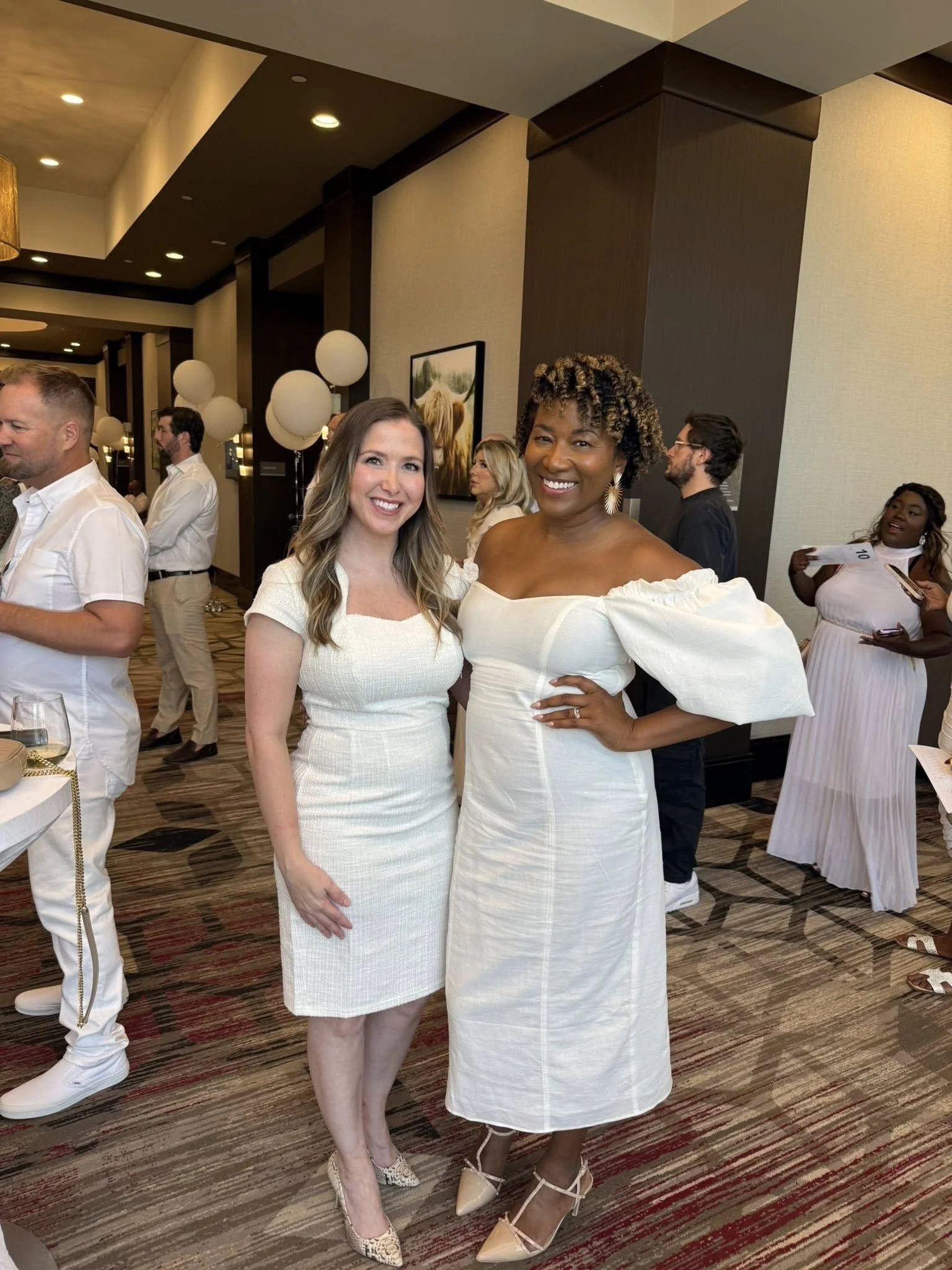 Two women smiling and posing for a photo at a lively indoor event, dressed in white dresses, with other guests in the background.