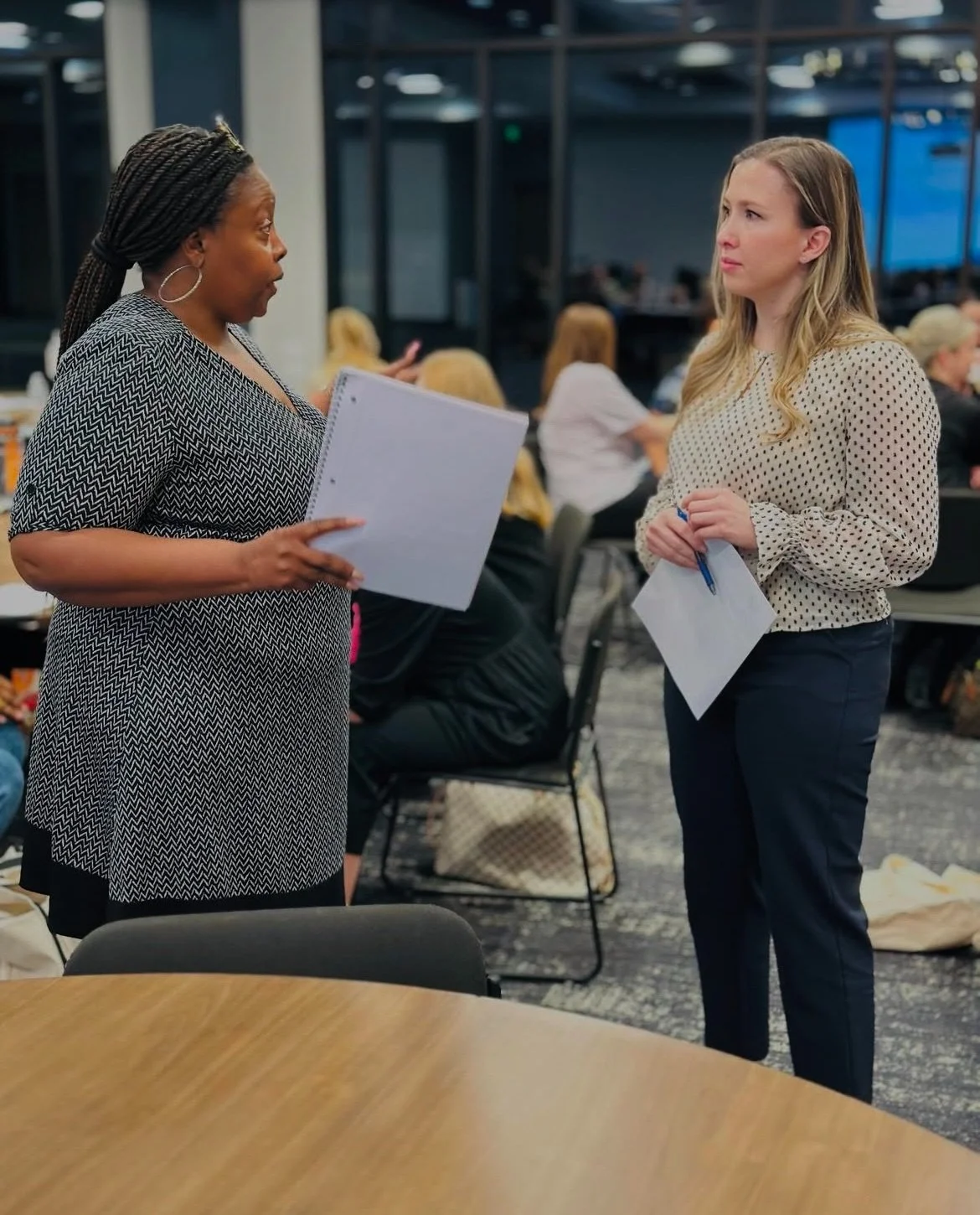 Two women having a discussion indoors, one holding a notepad and the other holding paper and a pen, with other people seated at tables in the background.