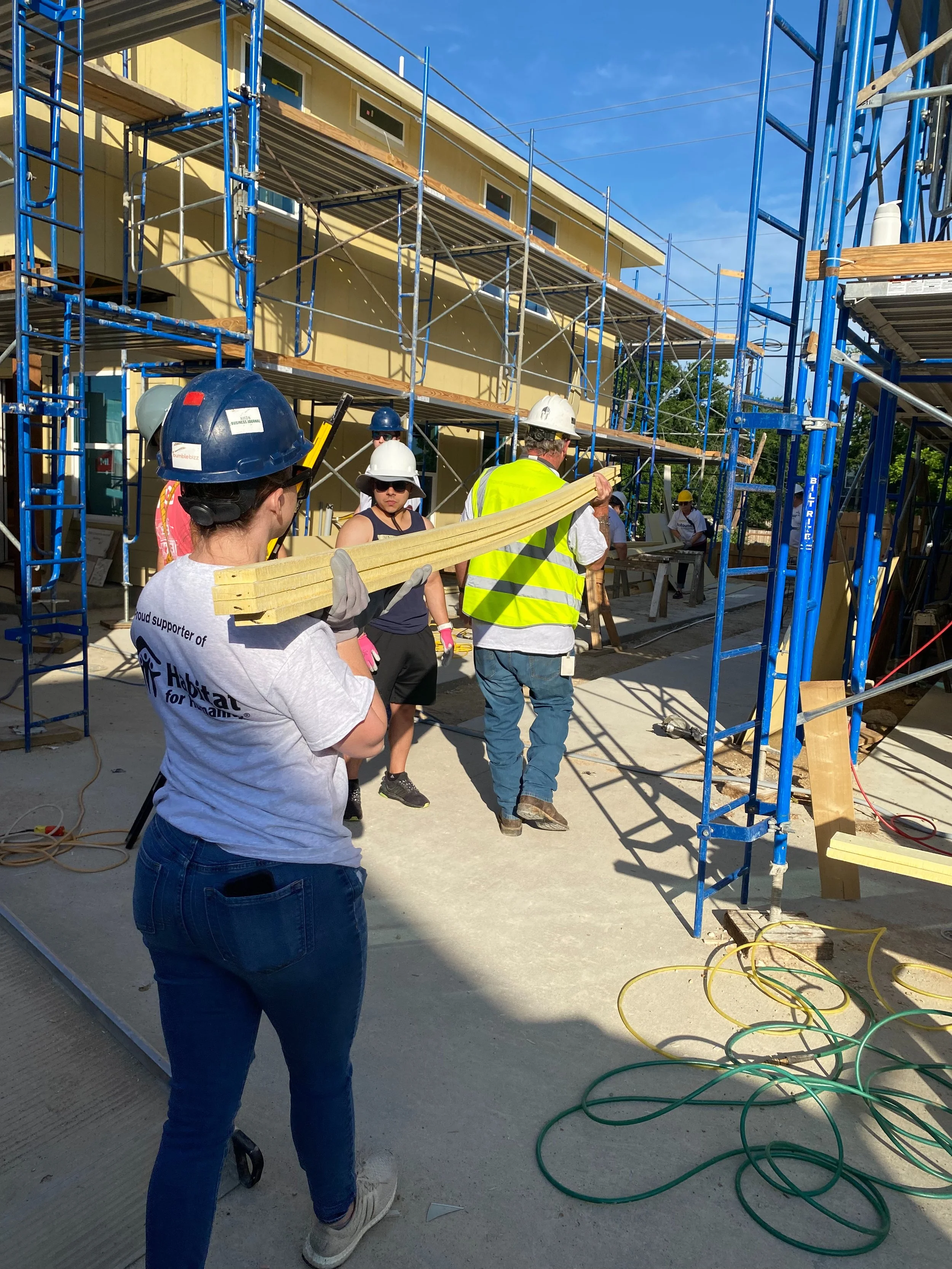 Construction workers on site, some wearing helmets and safety vests, working on building a structure with blue scaffolding and wood materials.