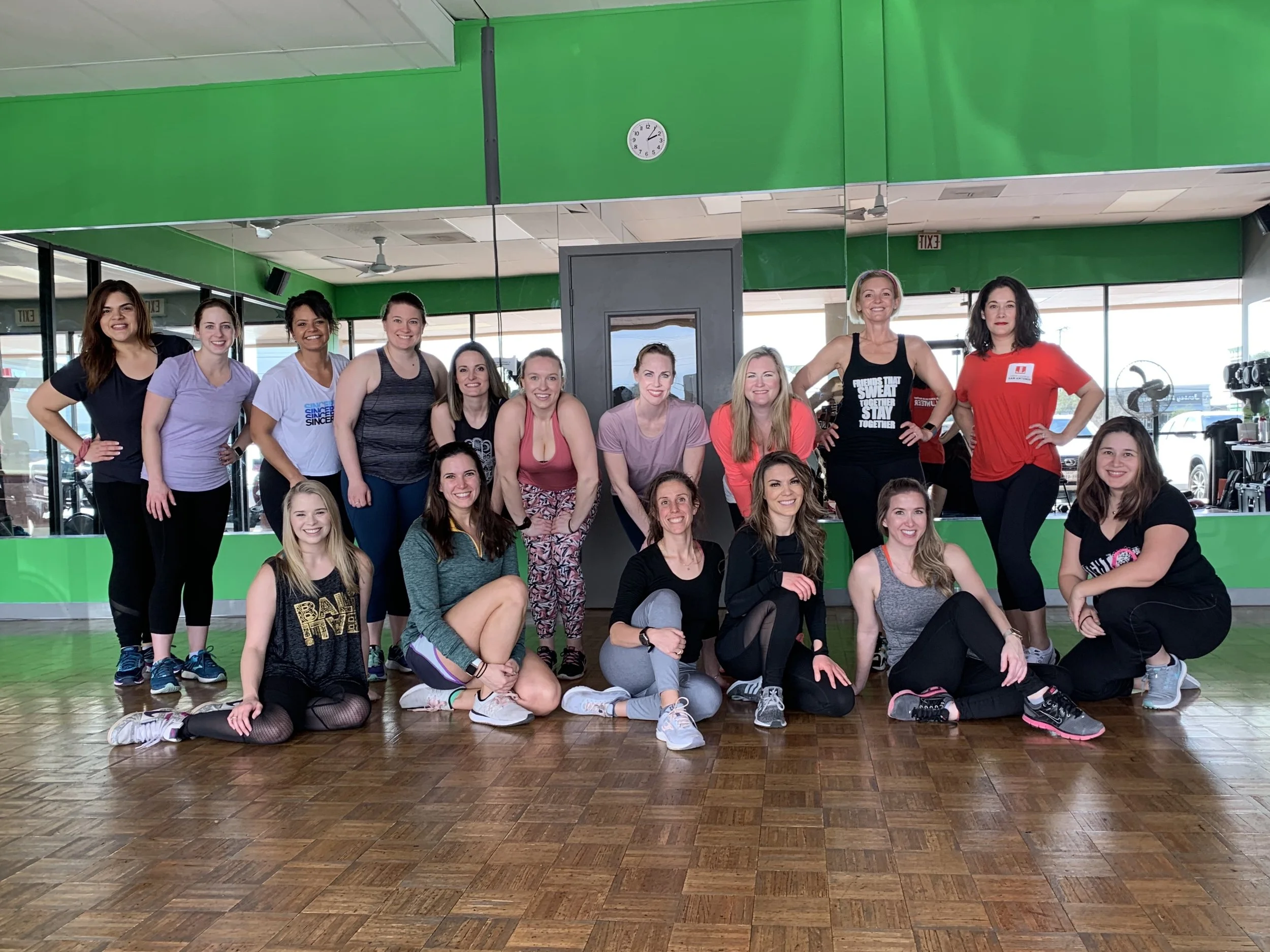 Group of women in workout clothes posing in a fitness studio with green walls and a wood floor.