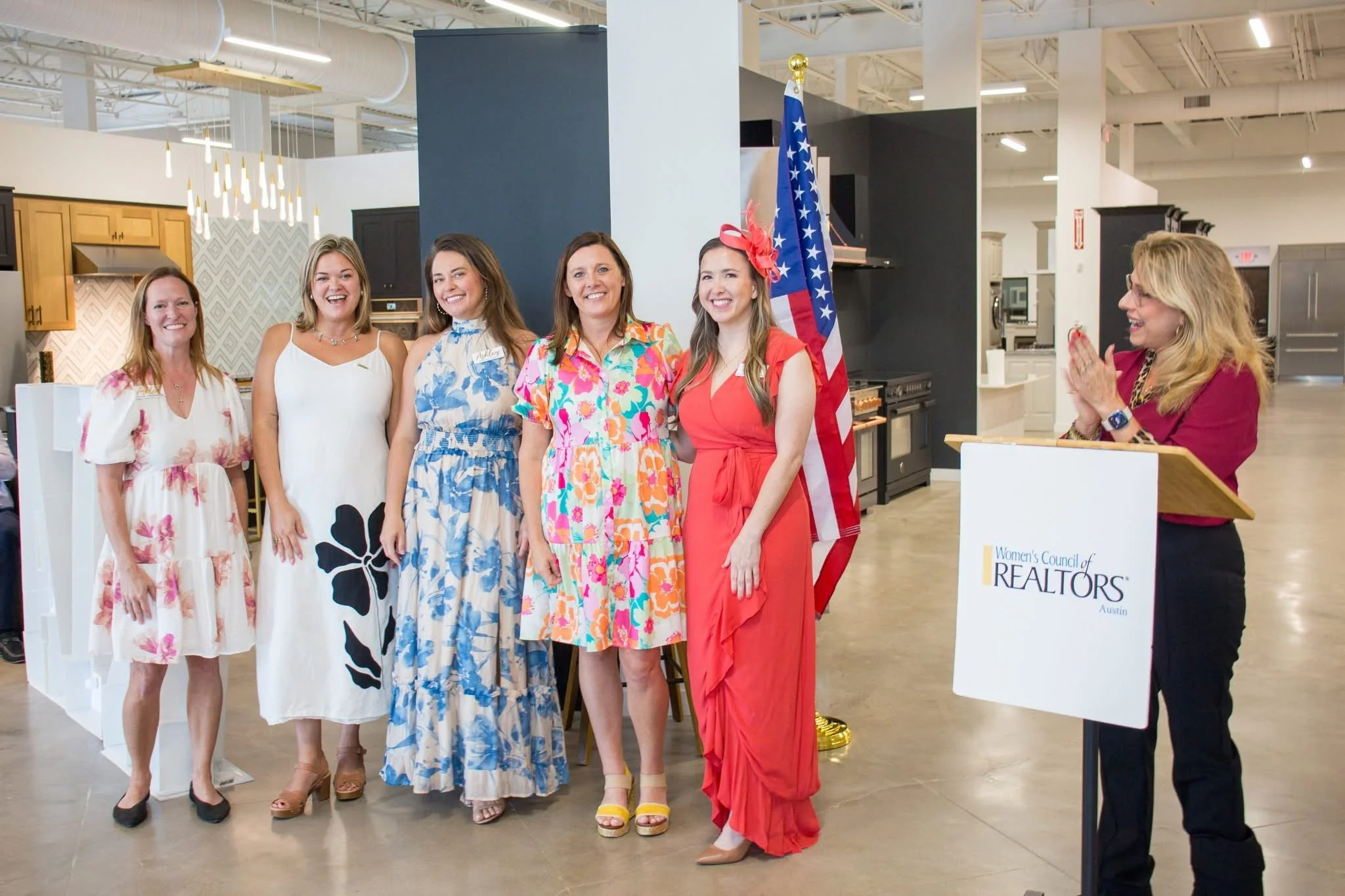 Six women in colorful dresses standing in a line at an indoor event, smiling and posing for the photo, with a blue column, American flag, and a woman at a podium with a Women's Council of REALTORS sign visible in the background.