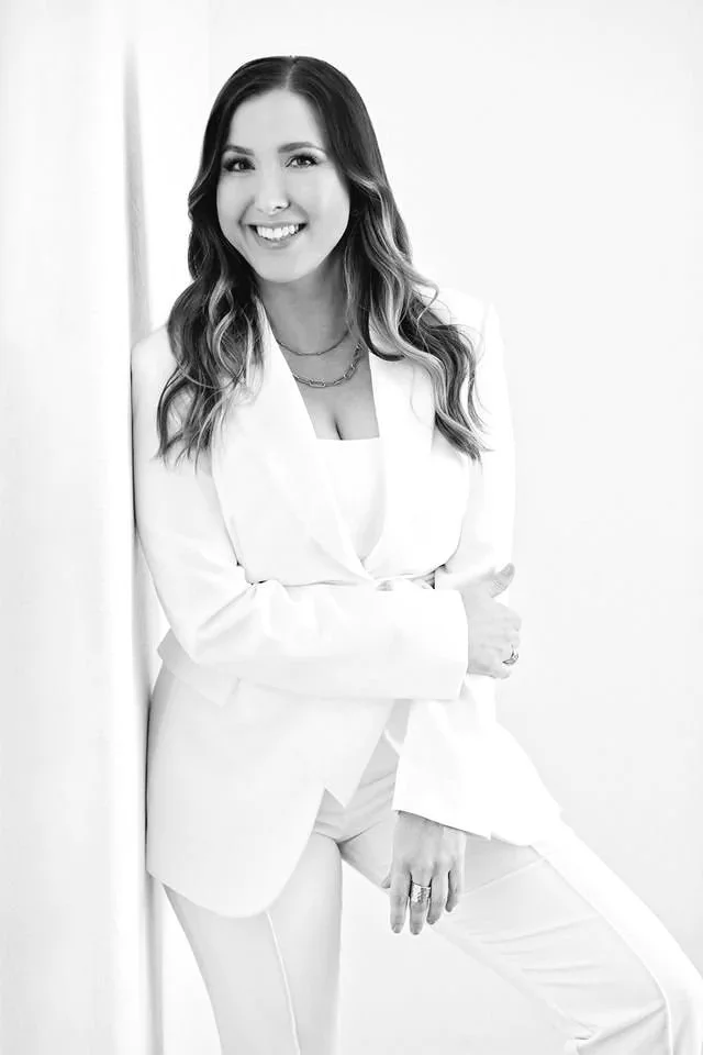 A woman with long wavy hair smiling, wearing a white blazer and matching pants, standing against a light background.