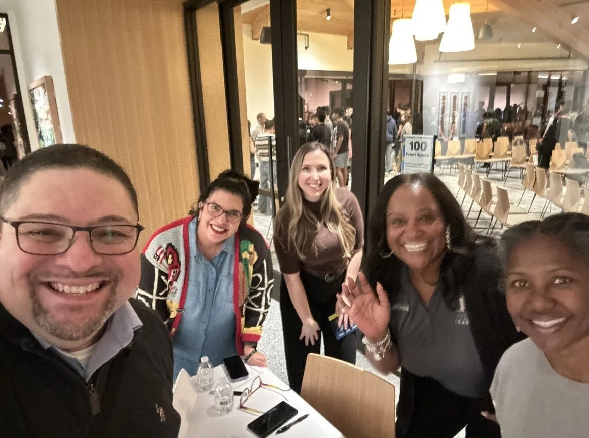 Group of five smiling women and men taking a selfie at a table during an event, with a glass wall and seated guests visible in the background.