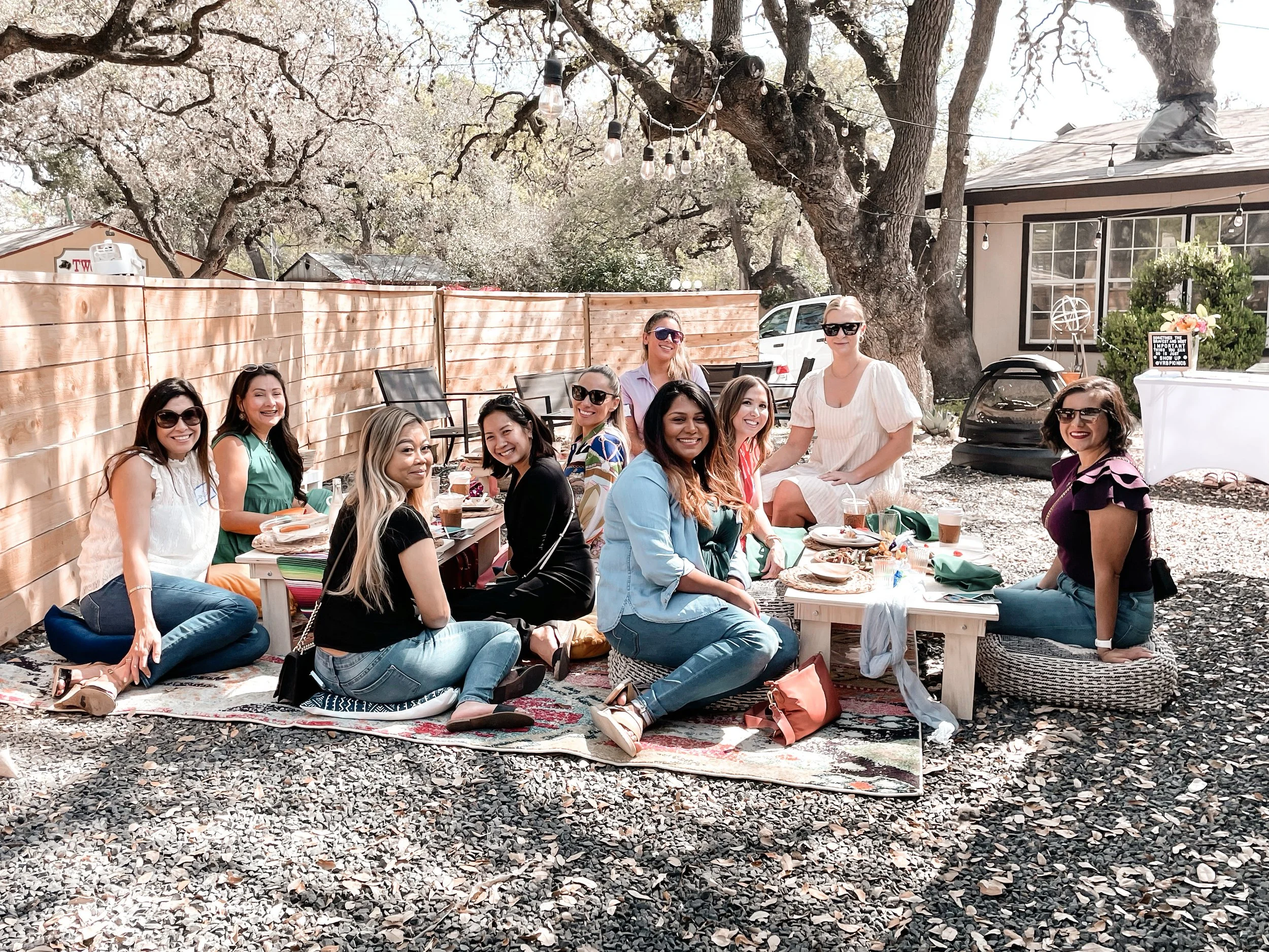 A group of ten women sitting on cushions and a low table outdoors under trees, enjoying a meal together in a backyard with string lights and a wooden fence.