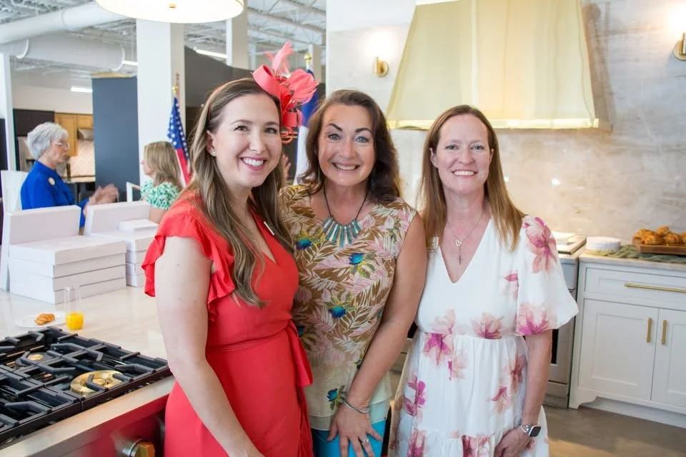 Three women smiling at an event, standing in a modern kitchen or bakery setting, dressed in colorful summer dresses.