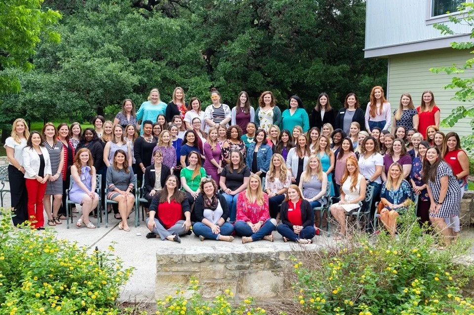 Group of women and girls gathered outdoors for a photo in front of trees and a building, some seated and some standing.
