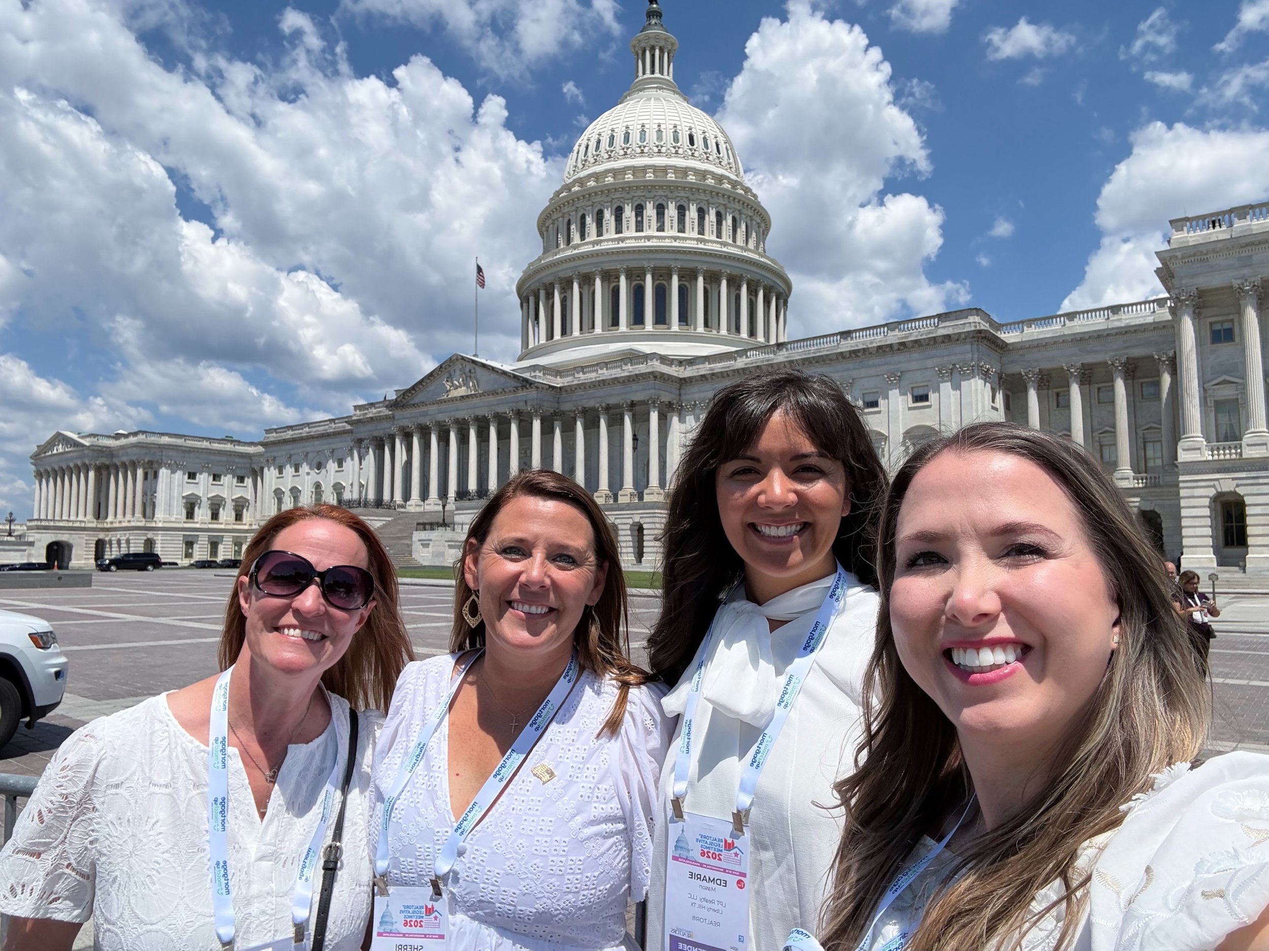 A group of five women smiling in front of the United States Capitol building on a sunny day with partly cloudy skies.