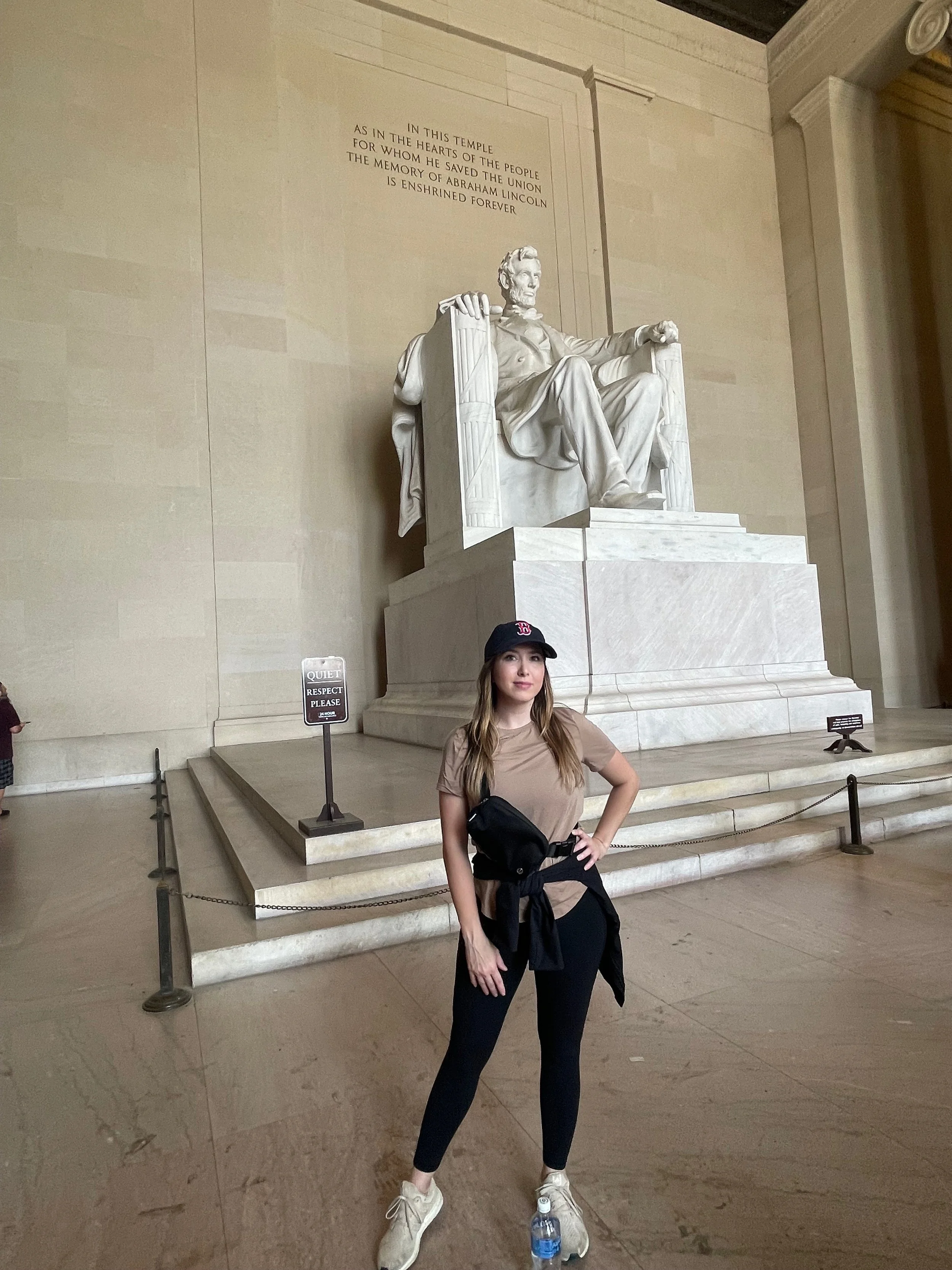 A woman in casual attire, wearing a Boston Red Sox cap, standing in front of the Lincoln Memorial in Washington, D.C. She has a black jacket tied around her waist and is wearing beige sneakers. A water bottle is on the floor near her. The Lincoln Mem