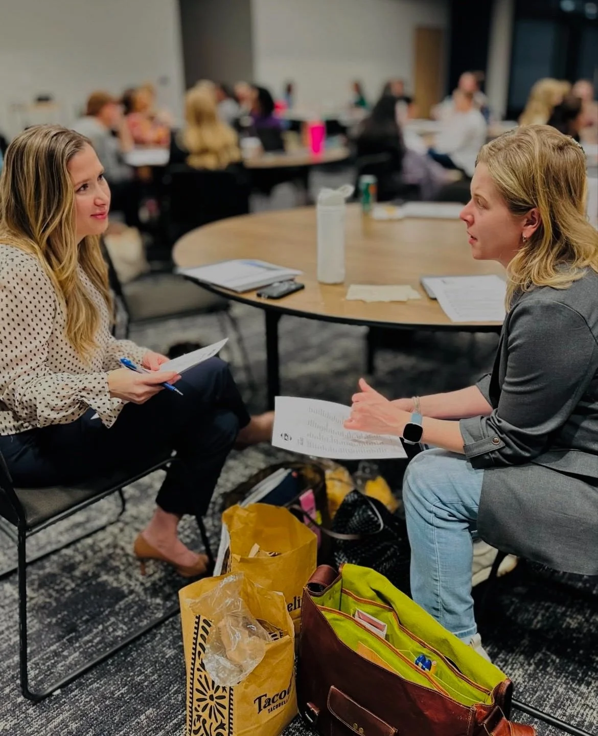 Two women sitting at a table engaged in a conversation during a conference or meeting, with notebooks, papers, and a bag on the floor.
