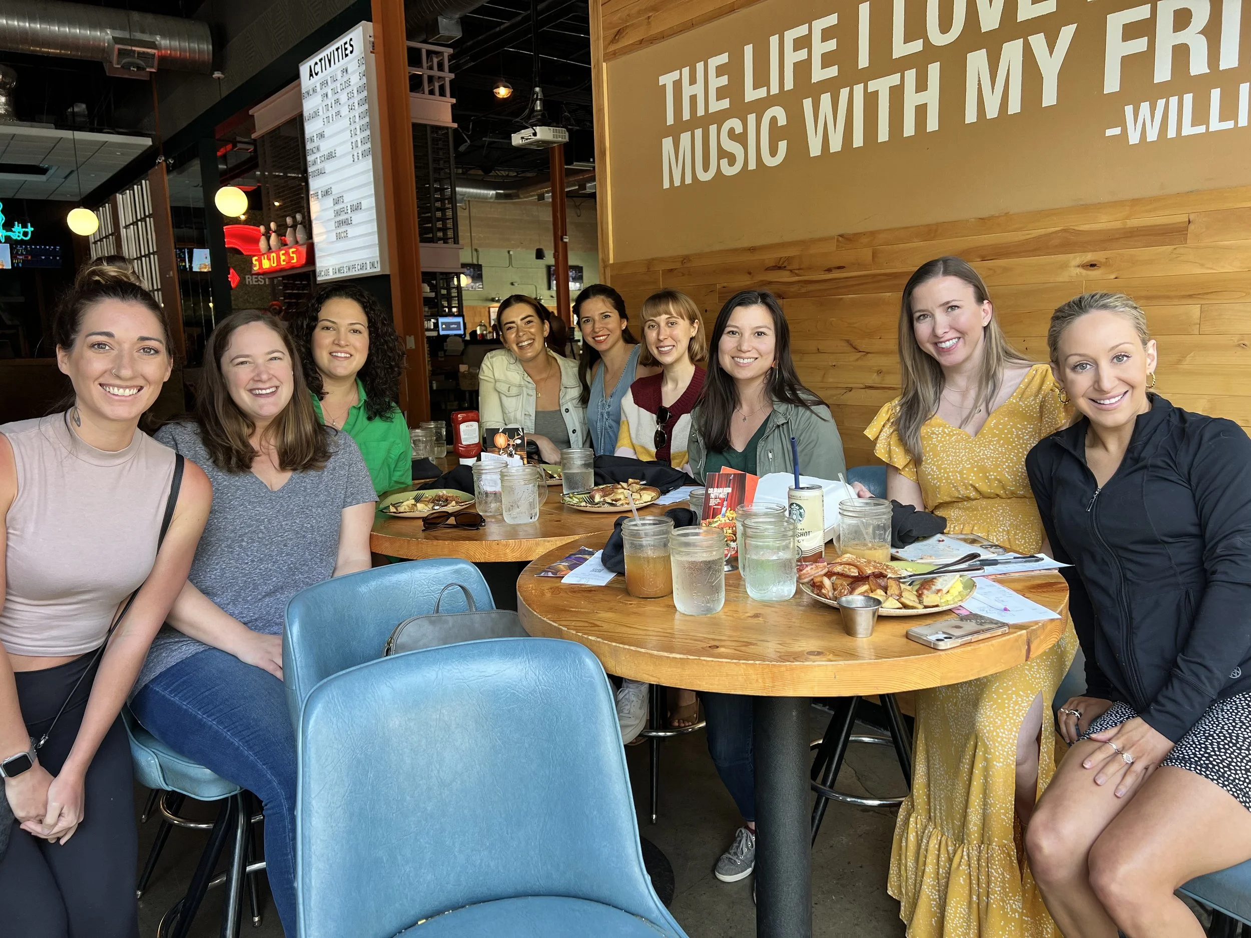 A group of nine women sitting around a table in a restaurant, smiling, with food and drinks in front of them. A large yellow wall with white text and wooden paneling in the background.
