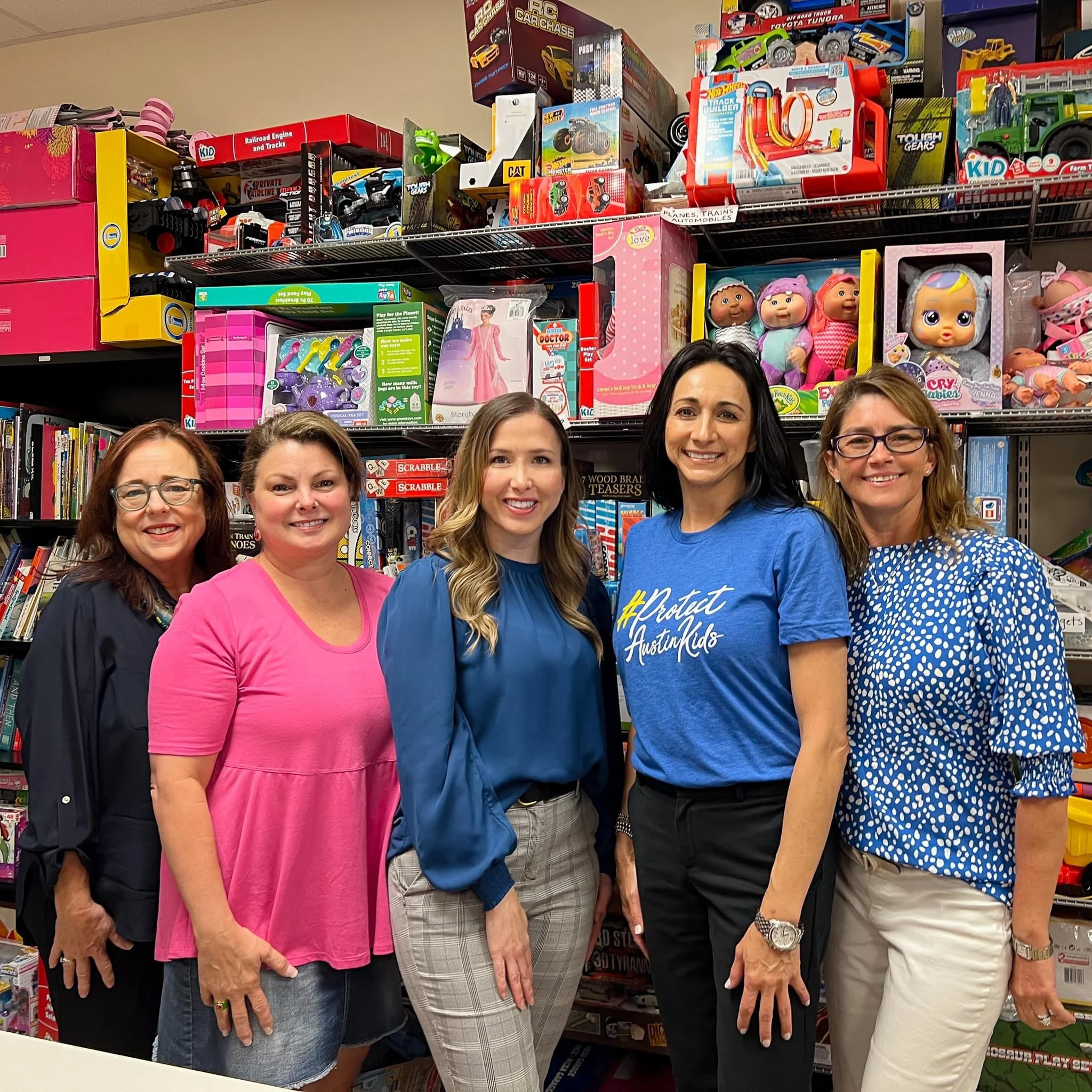 Five women standing in front of shelves filled with toys, including dolls, racing cars, and building sets, inside a store.