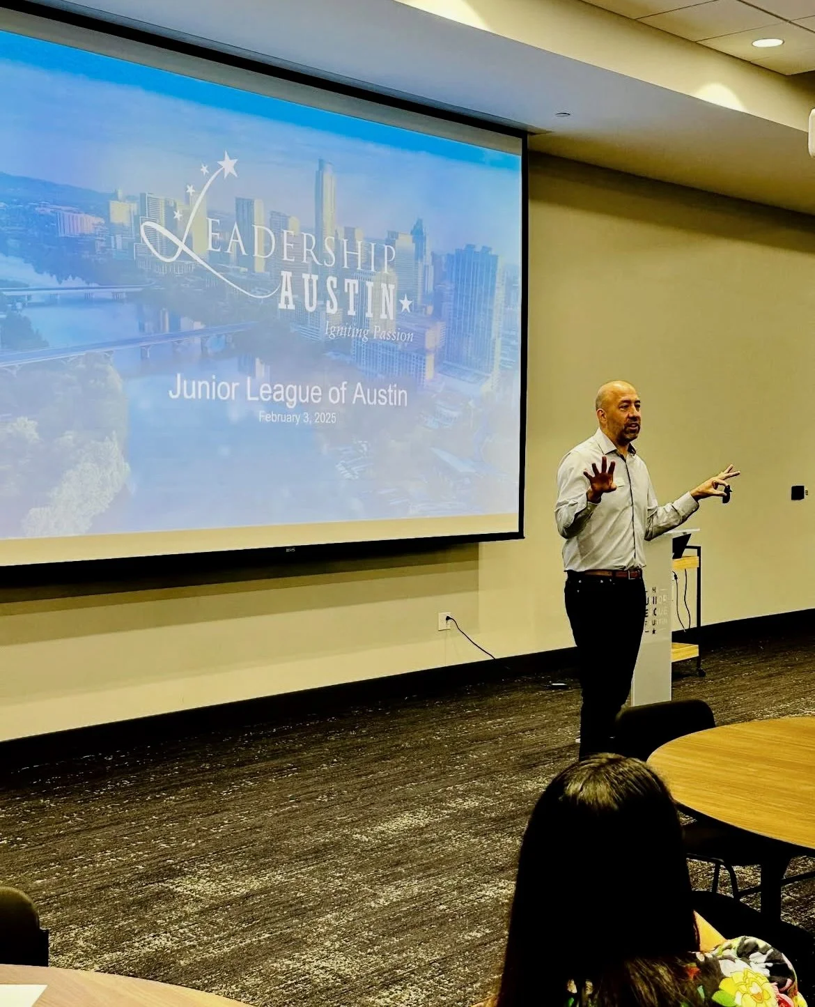 A man giving a presentation in a conference room with a screen behind him that reads 'Leadership Austin Igniting Passion Junior League of Austin February 3, 2025'. The presenter is standing near the wall, gesturing with both hands, and there are audi