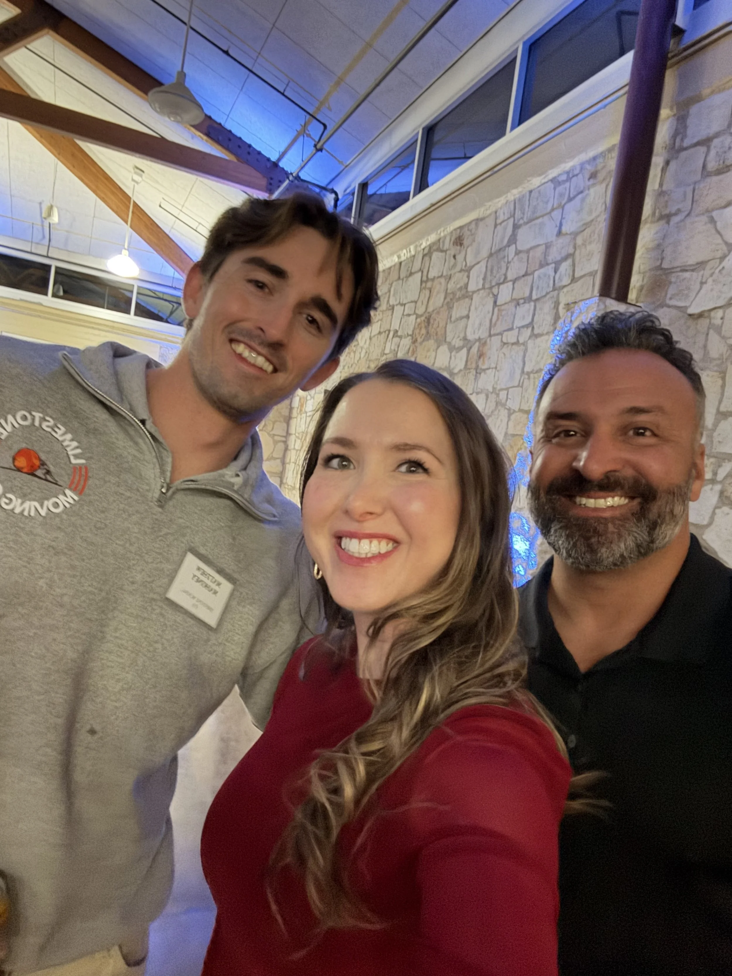 Group photo of three people smiling at a social event. The man on the left wears a gray hoodie with a logo and a name tag. The woman in the middle has long, wavy hair and wears a red top. The man on the right has short hair, a beard, and wears a blac