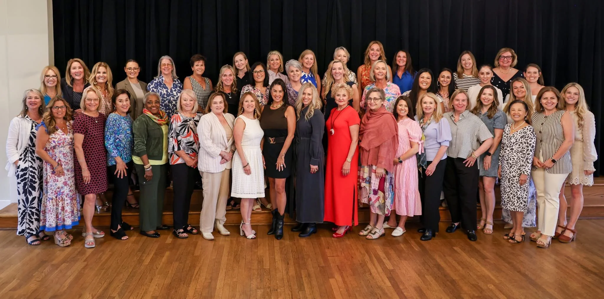 Group photo of many women standing on a wooden floor stage in front of a black curtain. They are smiling and dressed in colorful and formal attire.
