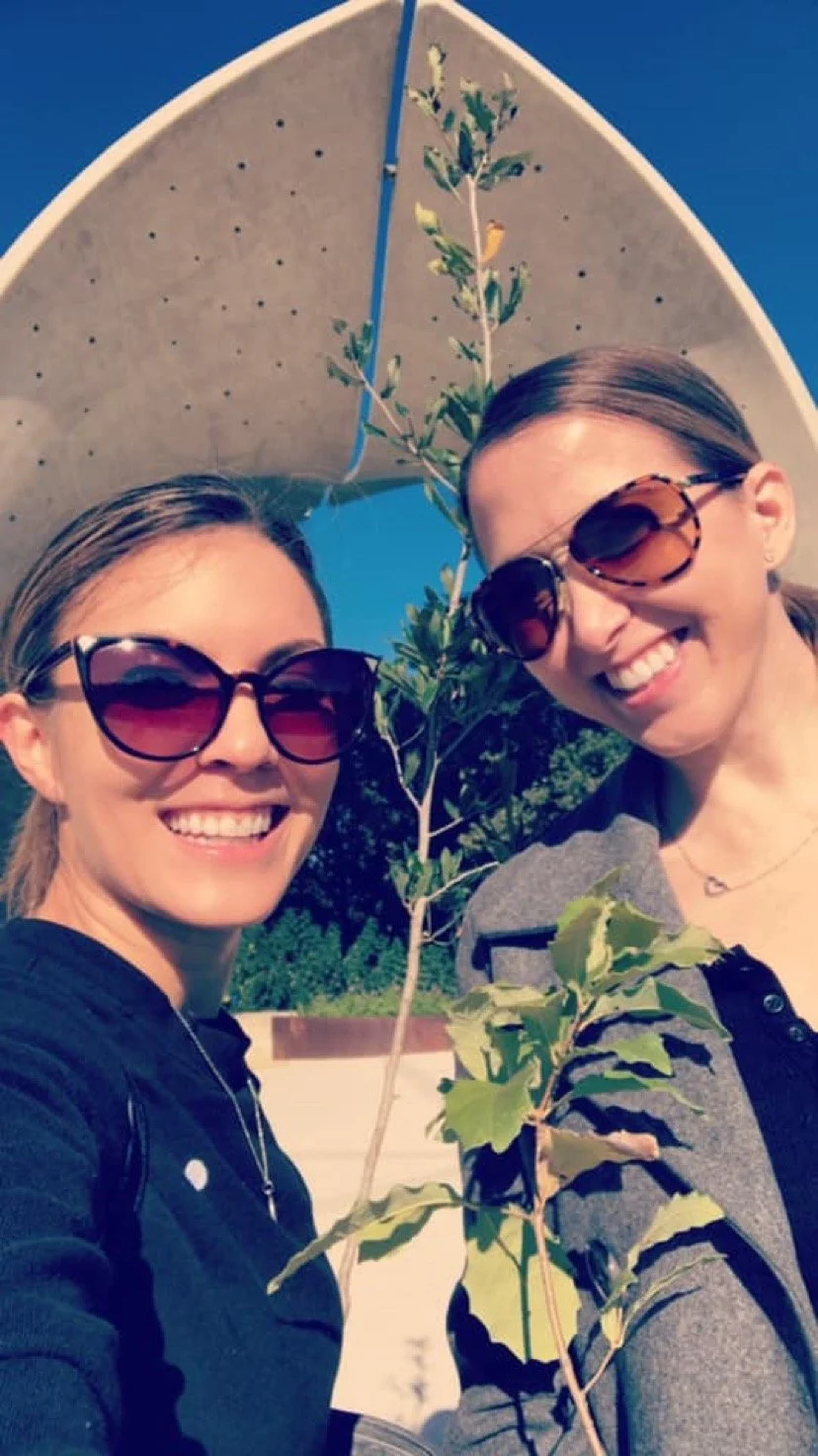 Two women smiling and taking a selfie outdoors under a modern, curved architectural structure, each holding small leafy branches, wearing sunglasses, with clear blue sky in the background.