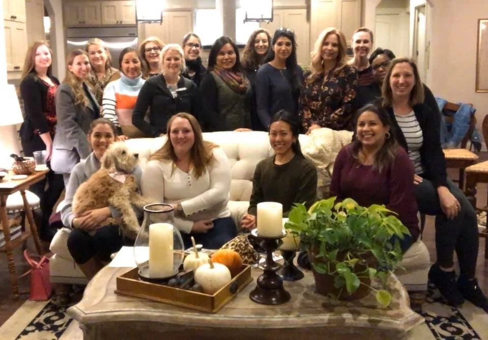 Group of women gathered in a living room, posing for a photo, with some smiling at the camera, and a dog sitting on a woman's lap. The room is decorated with candles, pumpkins, and a potted plant on a coffee table.