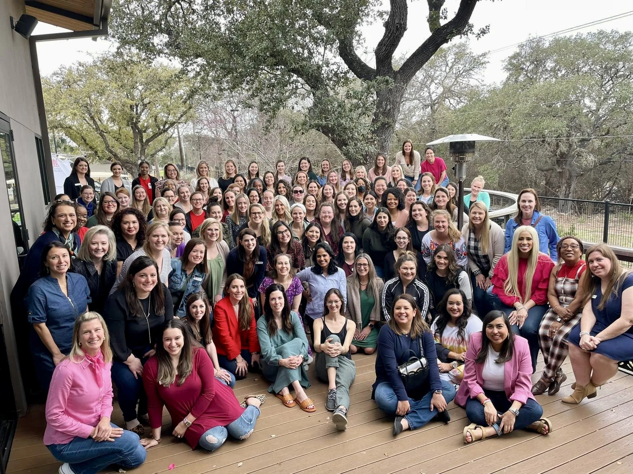A large group of women gathered on a wooden deck outdoors, smiling for a group photo with trees and a patio heater in the background.