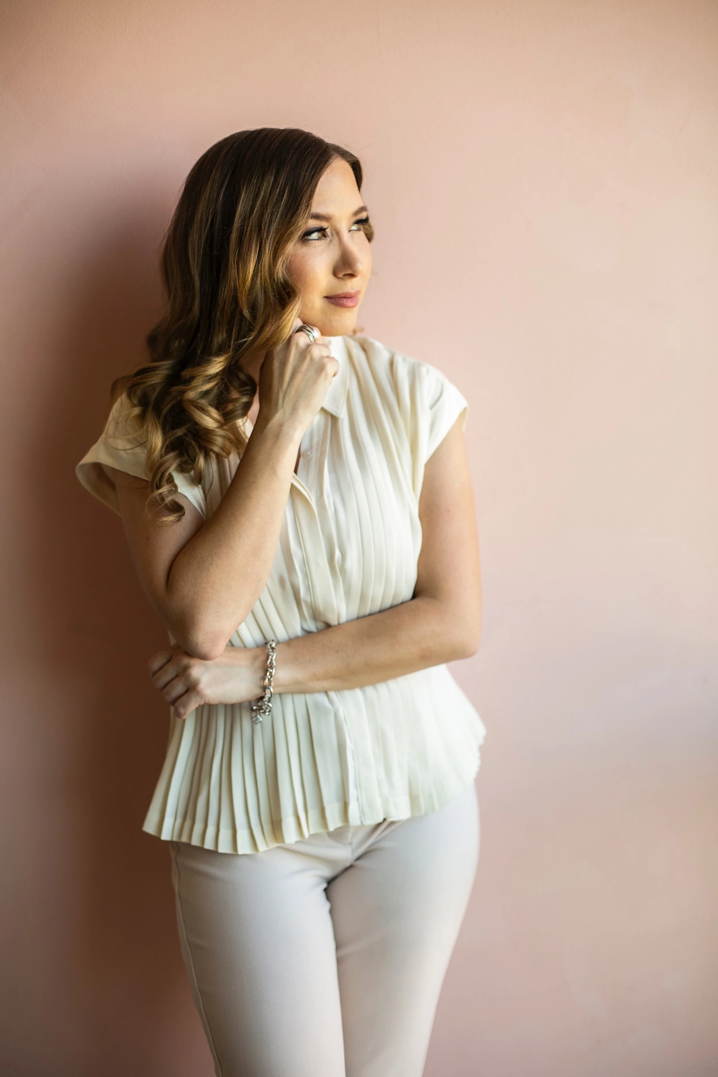 A woman with long, wavy brown hair, wearing a cream-colored pleated blouse and matching white pants, standing against a pink wall, looking to her left with her hand resting on her chin and a slight smile.