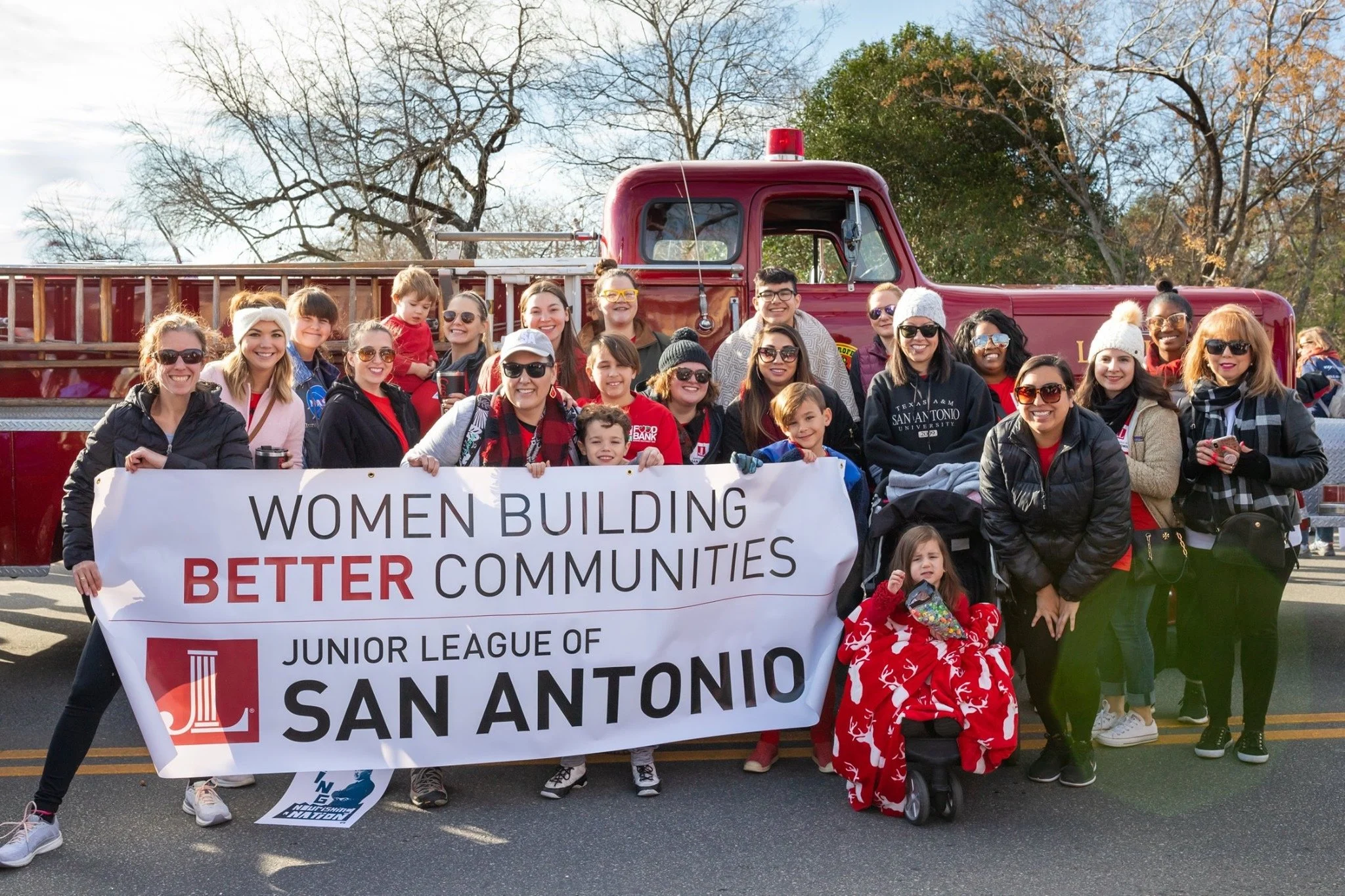 Group of people holding a banner that reads 'Women Building Better Communities,' standing in front of a red fire truck, with some children and adults smiling, during a parade or community event.