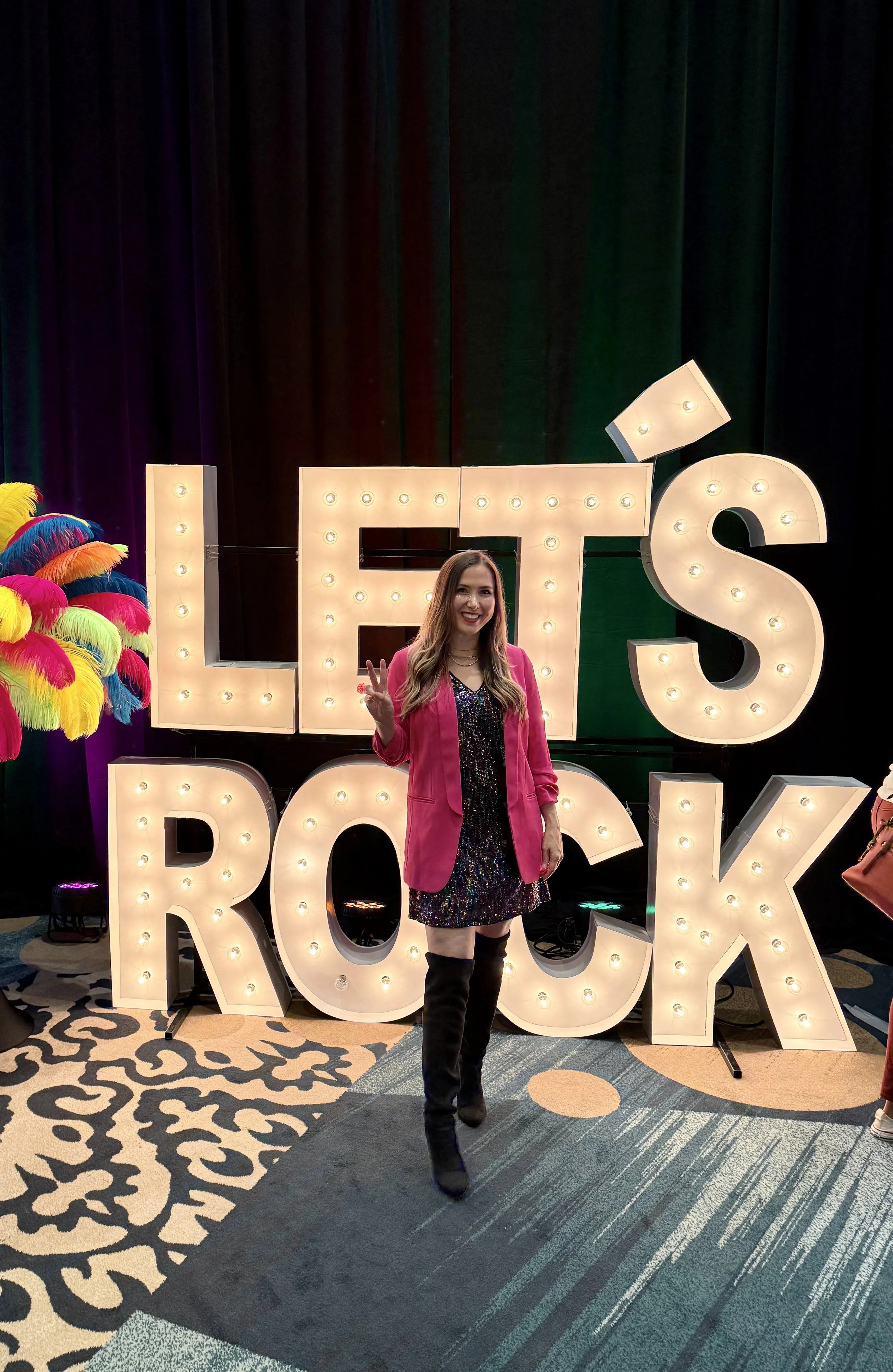 April Guerra in pink blazer and black dress standing in front of large illuminated letters spelling 'LET'S ROCK', making a peace sign. Colorful feather decoration on the left, dark stage curtain background, patterned carpet on the floor.